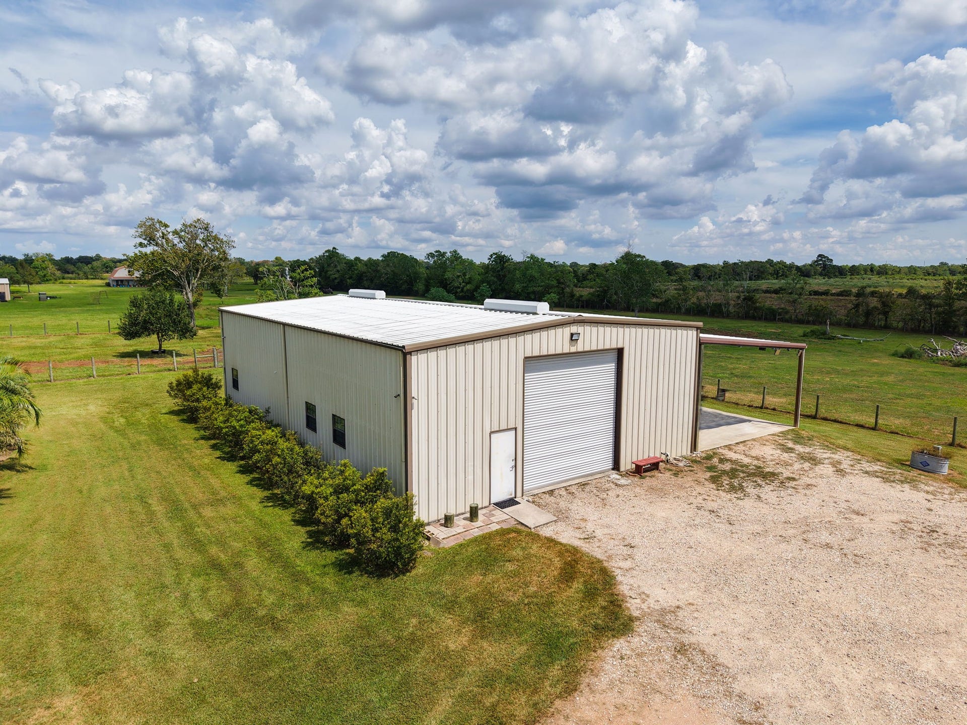 Expansive metal workshop with a large roll-up door, set on a spacious, fenced property surrounded by lush greenery and open skies.