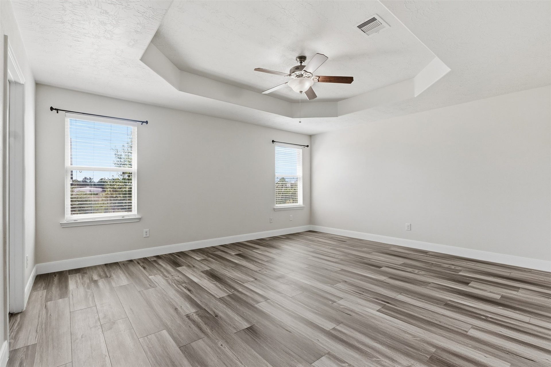 Sizable primary bedroom featuring wood-look flooring, neutral finishes, and a raised tray ceiling with a ceiling fan and light fixture. Multiple windows provide abundant natural light, while the generous room dimensions offer ample space for a variety of bedroom furniture arrangements.