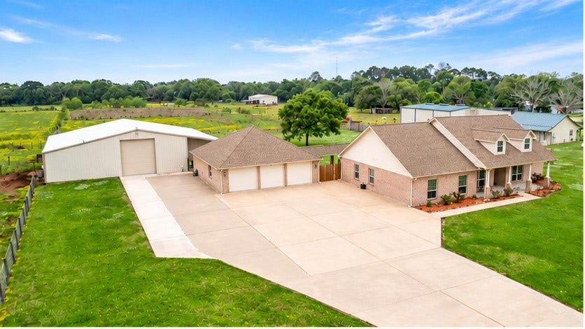 Impressive aerial view showcasing the brick home, spacious three-car garage, large workshop, and expansive concrete driveway, all surrounded by beautiful acreage and scenic country views.