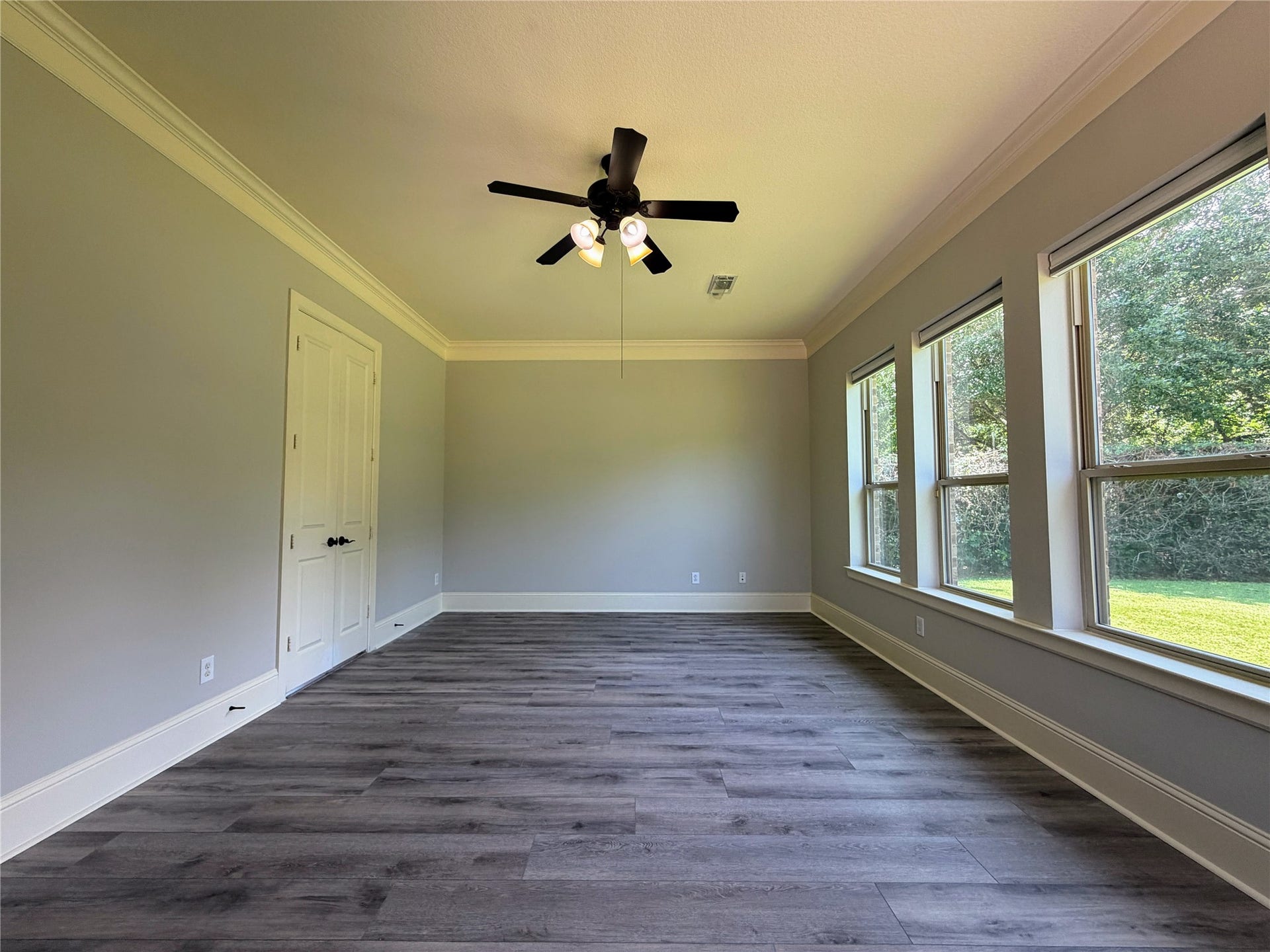 5119 Water Oak Crescent, Fulshear, TX 77441 Serene primary suite retreat! This spacious bedroom boasts stylish, modern wood-look flooring, elegant crown molding, a classic ceiling fan, and a wall of large windows overlooking the lush, private backyard space.