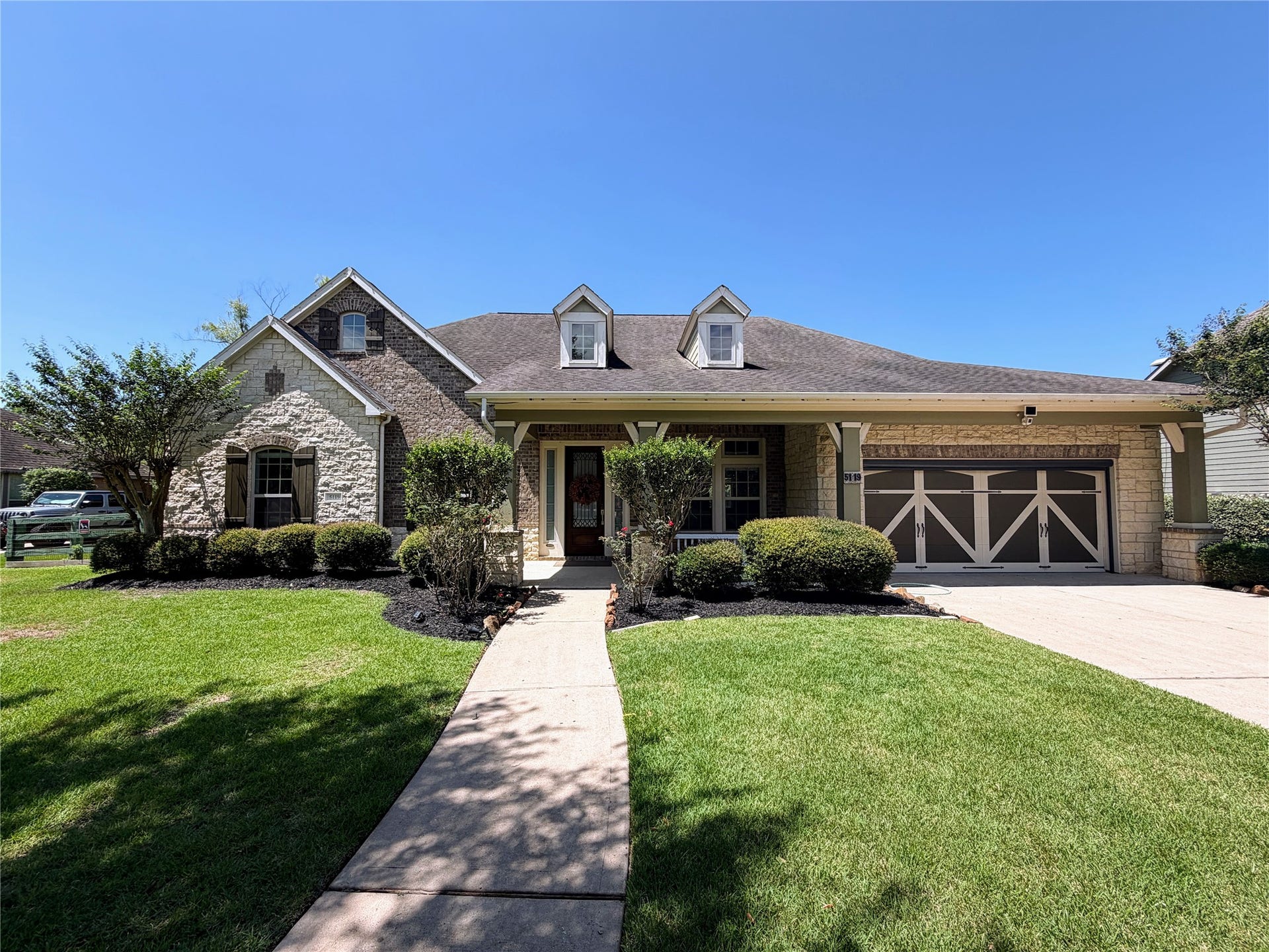 5119 Water Oak Crescent, Fulshear, TX 77441 Picture-perfect curb appeal! This charming 1.5-story estate showcases timeless stone and brick accents, beautifully manicured landscaping, a welcoming front porch, and an elegant garage door under a bright Texas sky.