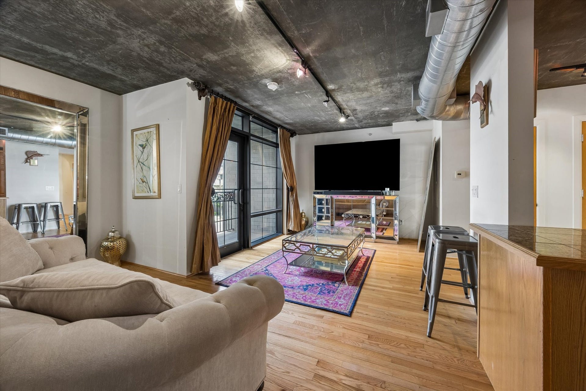 The living room offers a comfortable setting with generous natural light, hardwood flooring, and exposed concrete ceilings. Large glass doors and an open floor plan create an airy atmosphere throughout.