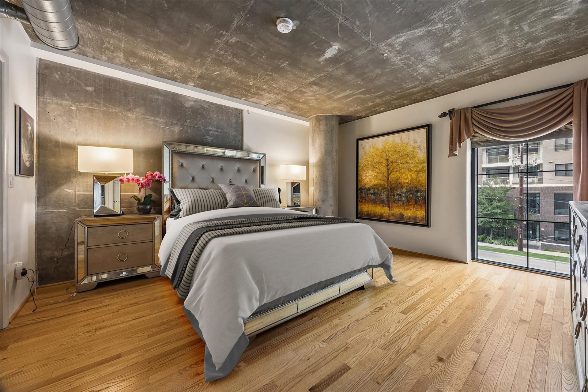The primary bedroom offers a spacious retreat with hardwood flooring, exposed concrete ceilings, and generous natural light. Its industrial loft character is balanced by warm finishes and ample room for comfortable furnishings.