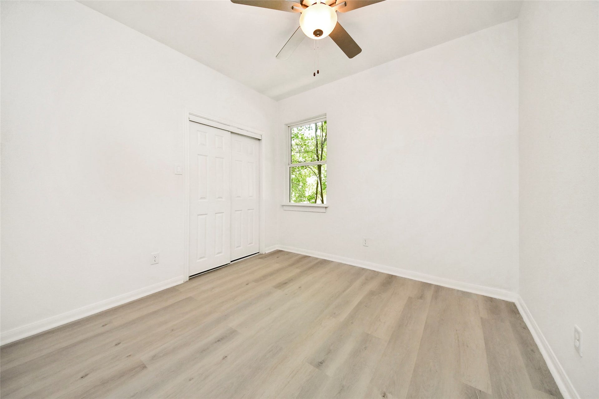 4615 Collingsworth Street, #AB, Houston, TX 77026 Bright and airy bedroom featuring sleek laminate flooring, a ceiling fan, and a closet with sliding doors, complemented by a large window offering a view of lush greenery.