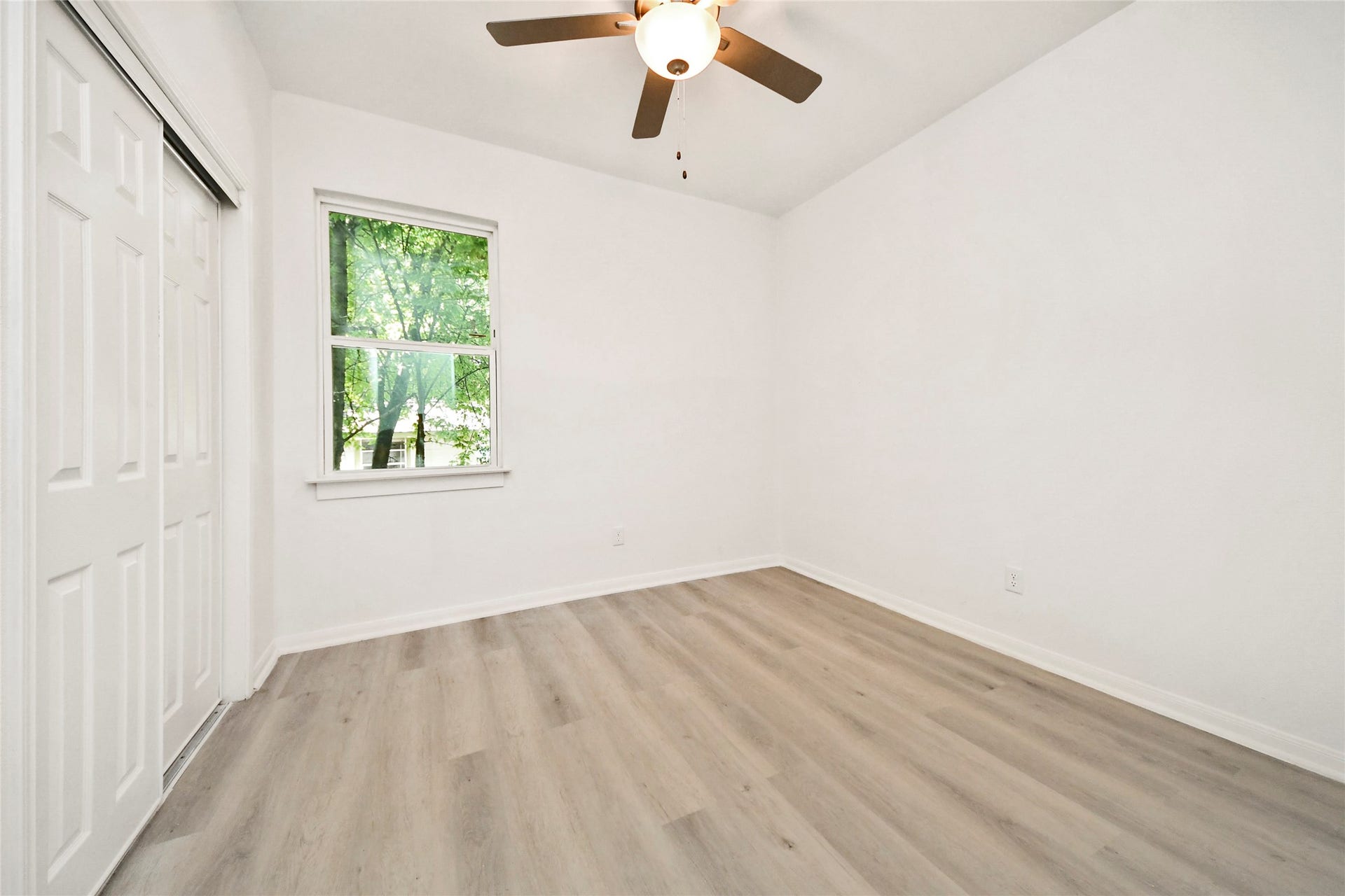 4615 Collingsworth Street, #AB, Houston, TX 77026 Bright and airy bedroom featuring sleek laminate flooring, fresh white walls, a ceiling fan, and a large window offering serene views of lush greenery.