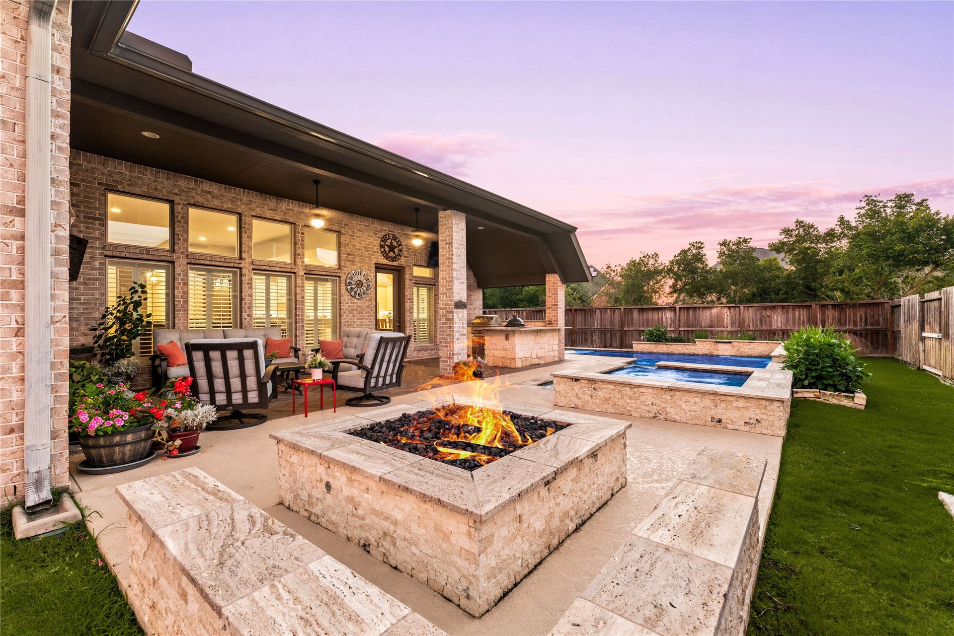 4103 August Light Court, Fulshear, TX 77441 Unwind under painted twilight skies in this spectacular resort-style backyard oasis.A massive, custom-built stone fire pit serves as the ultimate open-air gathering spot, surrounded by sprawling stone tile decking that extends effortlessly across the yard. In the background, a sparkling plunge pool and an integrated hot tub are framed by clean stone masonry and manicured green lawn spaces.