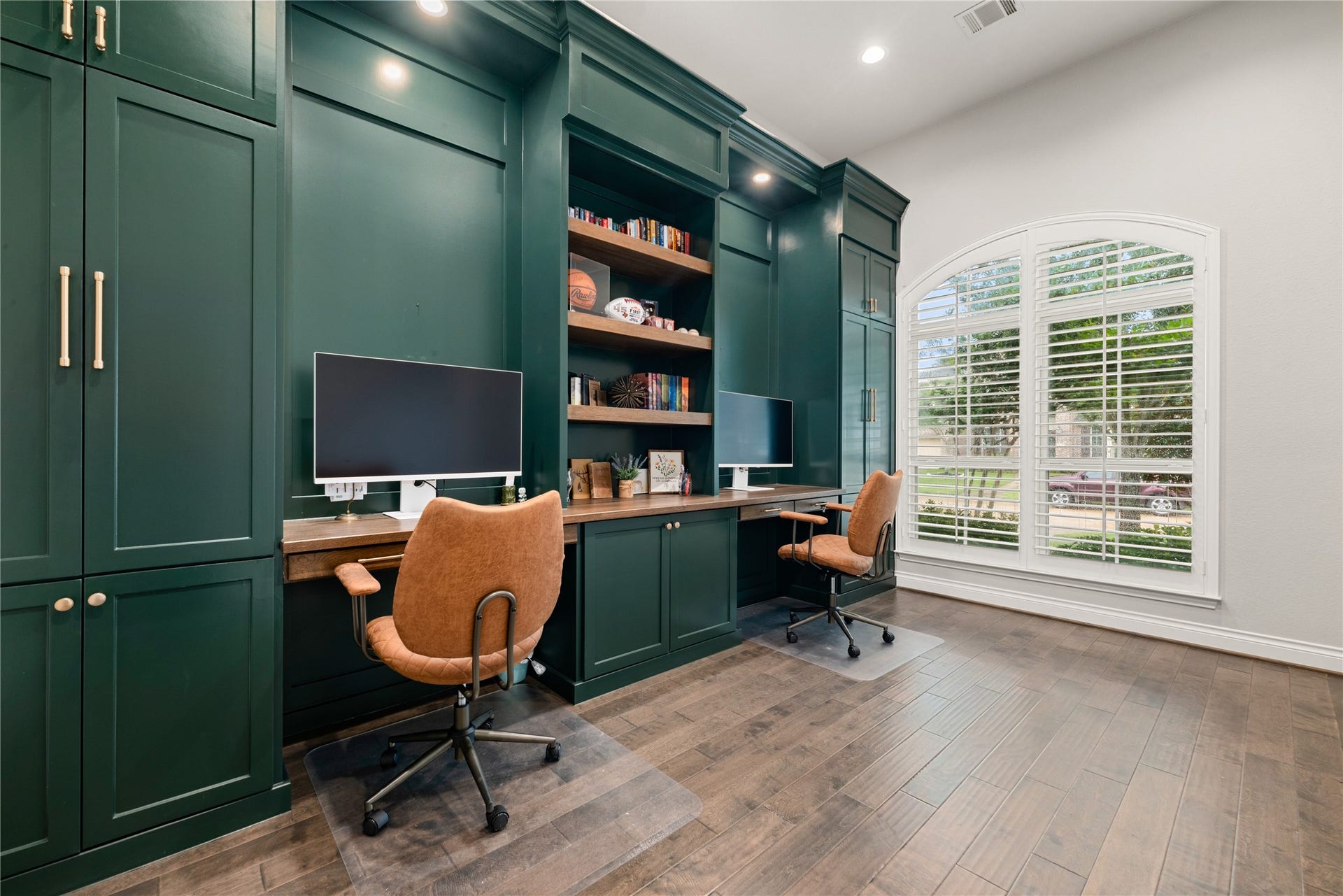 4103 August Light Court, Fulshear, TX 77441 The centerpiece of this sophisticated space is a massive, floor-to-ceiling bank of custom cabinetry painted in a rich, dramatic emerald green. This masterfully crafted unit integrates dual distinct workspaces featuring warm wood-toned desktop surfaces, expansive open display shelving, and an abundance of hidden storage. Modern satin-gold hardware punctuates the cabinetry, adding a refined, high-end touch to the executive design.