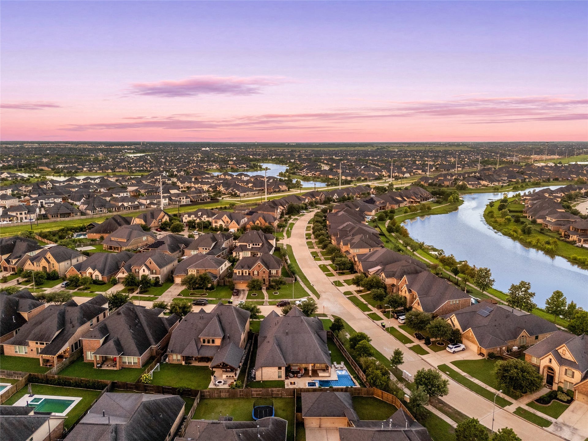 4103 August Light Court, Fulshear, TX 77441 This commanding aerial view showcases a meticulously designed neighborhood layout, beautifully bordered by a winding, scenic waterway. Tree-lined streets and wide sidewalks curve gracefully through the development, connecting rows of stunning executive-level estates characterized by clean masonry lines and dramatic rooflines.
