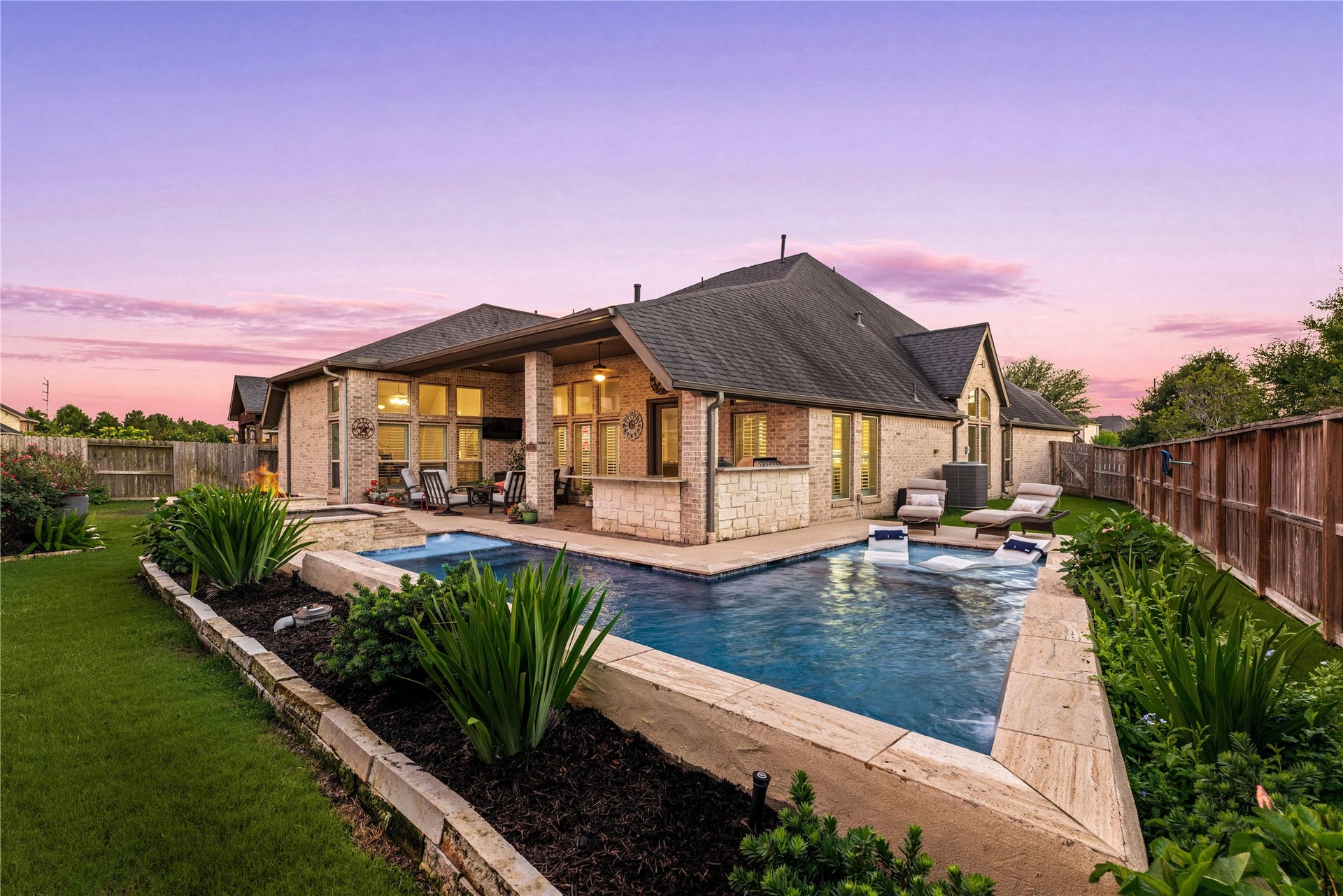 4103 August Light Court, Fulshear, TX 77441 From this wide panoramic view, the entire estate shines beneath a twilight sky, showcasing the vast covered entertaining pavilion, stone outdoor kitchen, and warm interior lighting glowing through the extensive walls of windows.
