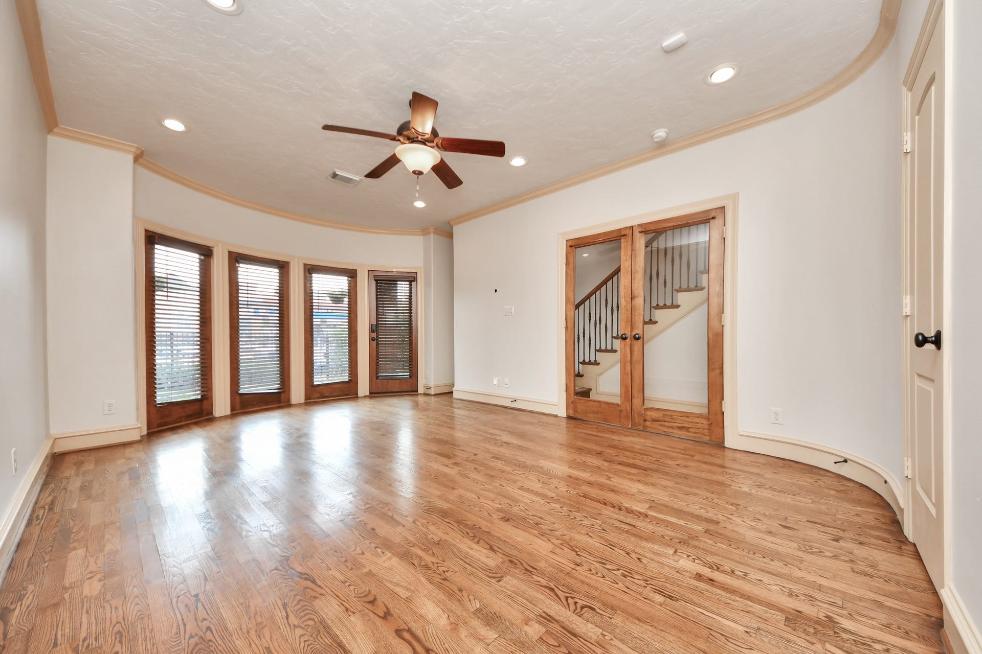 Elegant first floor bedroom/study area featuring rich hardwood floors, a stylish ceiling fan, and abundant natural light from large windows with wooden blinds. French doors open to a staircase, enhancing the home's charm.
