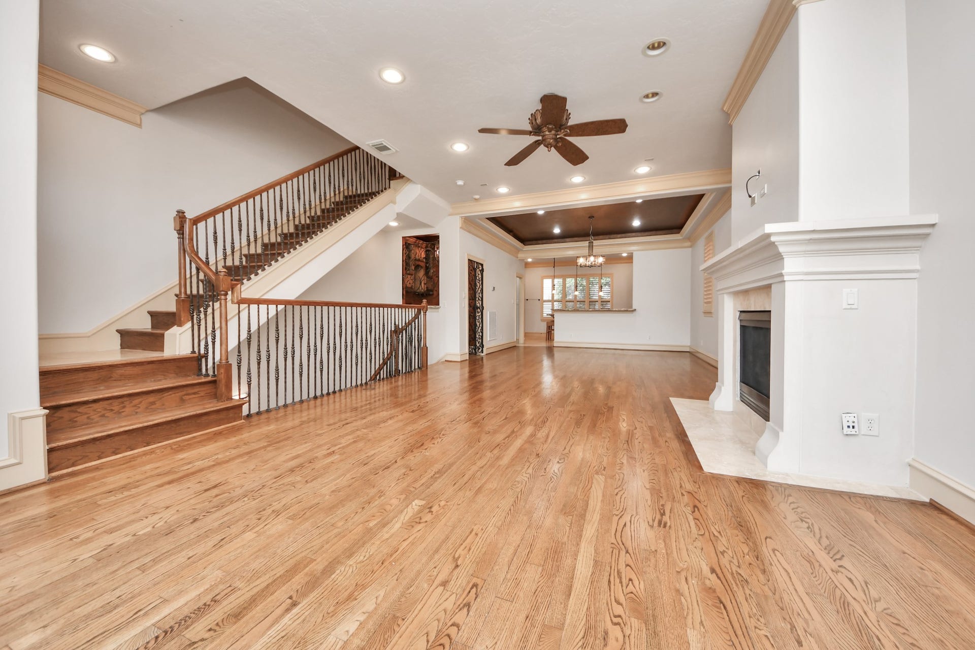 Spacious living area featuring elegant hardwood floors, a stylish coffered ceiling with recessed lighting, a cozy fireplace, and an open staircase with wrought iron railings.