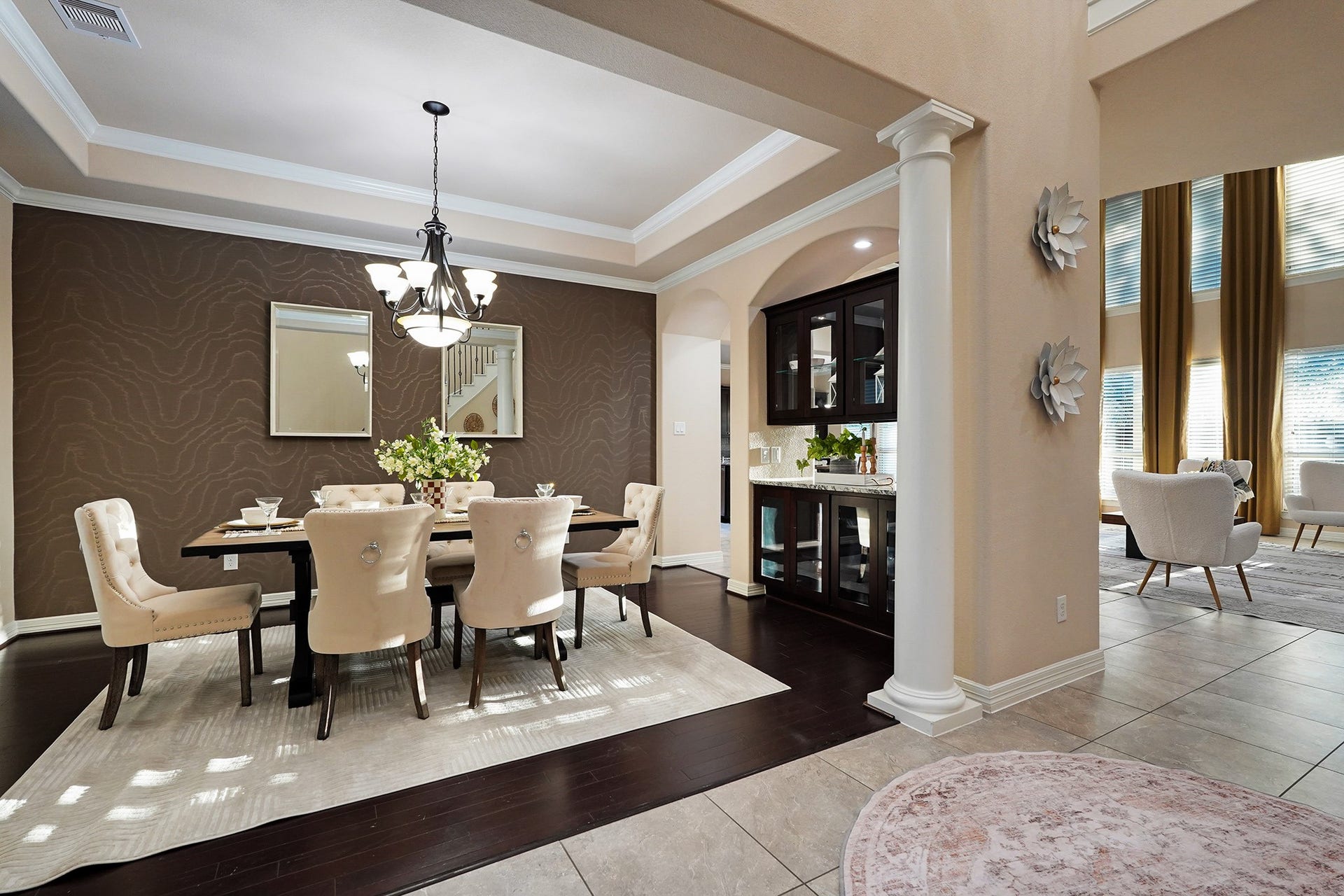 3102 Barrons Way, Sugar Land, TX 77479 This elegant dining area features a stylish chandelier, coffered ceiling, and a warm accent wall. It opens to a bright living space with tall windows and high ceilings, offering a sophisticated and inviting atmosphere.