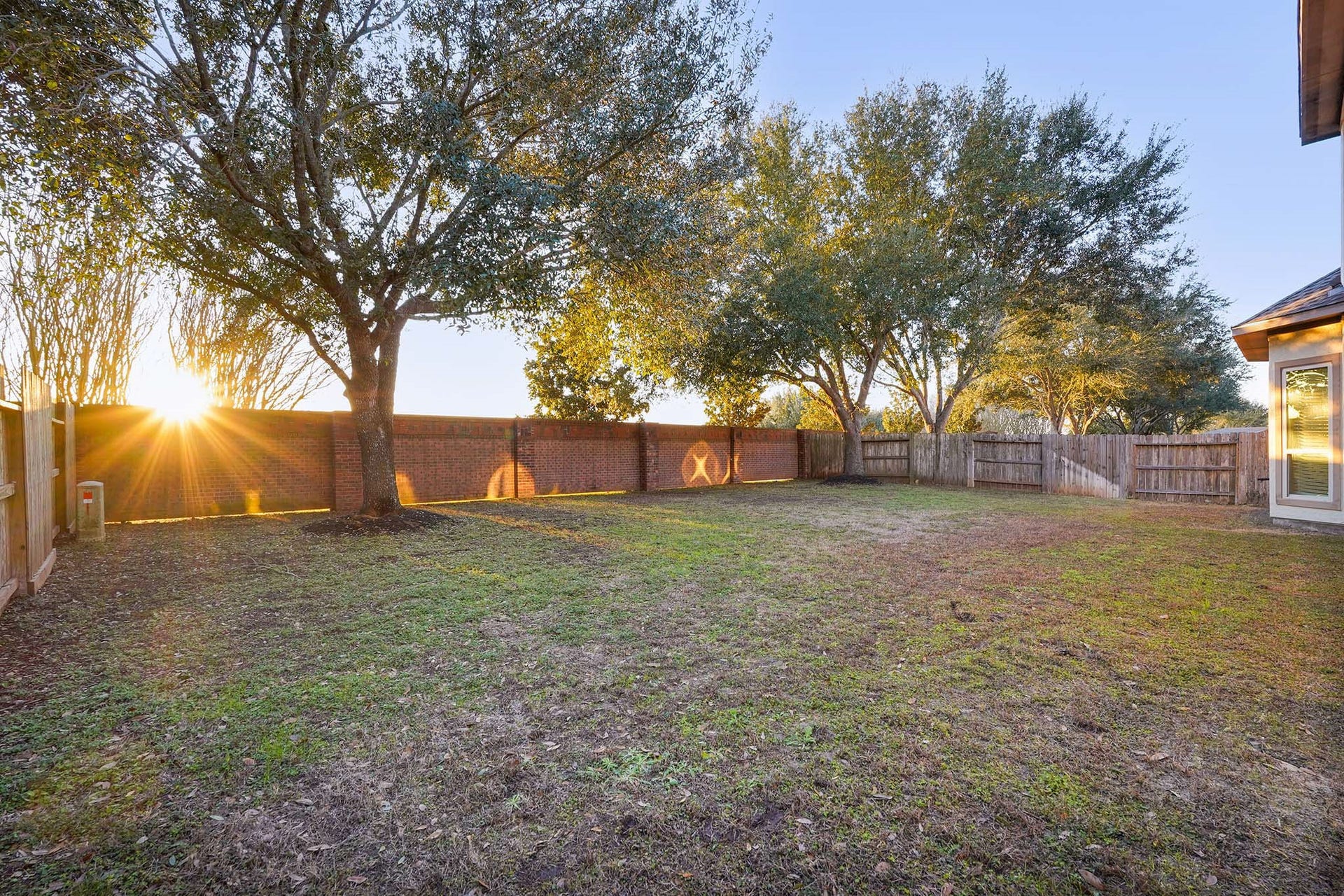 3102 Barrons Way, Sugar Land, TX 77479 The backyard bordered by a wooden and brick fence. Mature trees provide shade, and the setting sun casts a warm glow, creating a peaceful atmosphere.