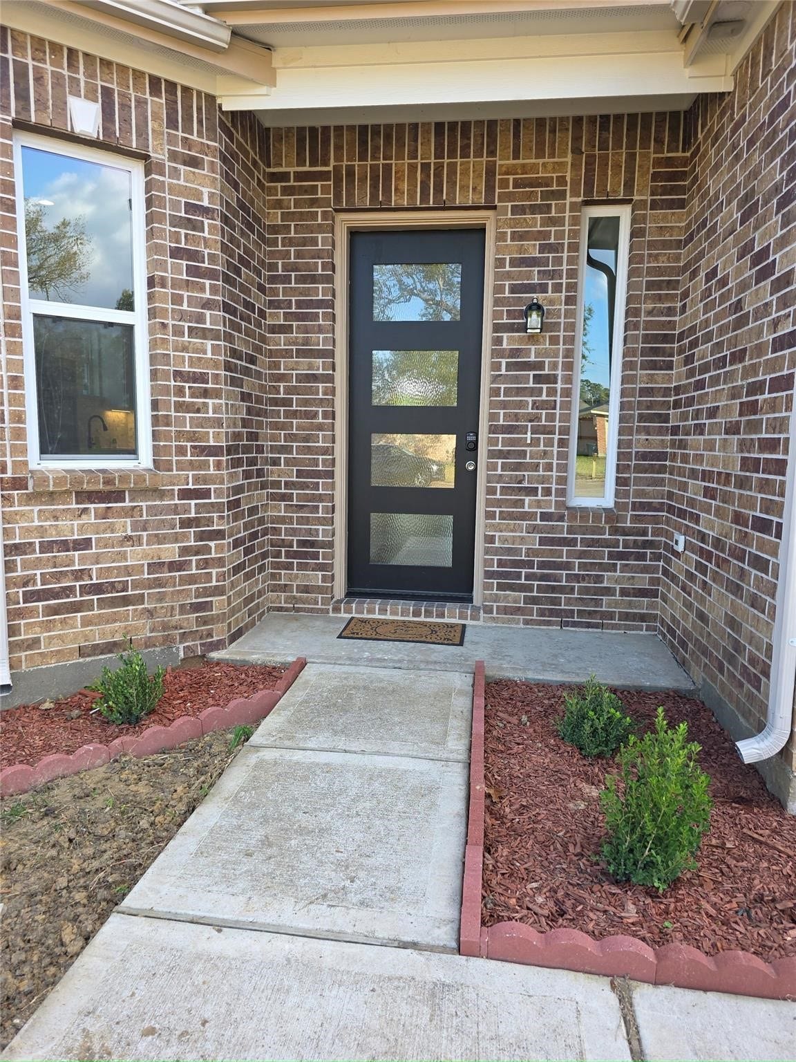 Charming brick entryway with a modern black door, framed by neatly landscaped flower beds and a welcoming path.