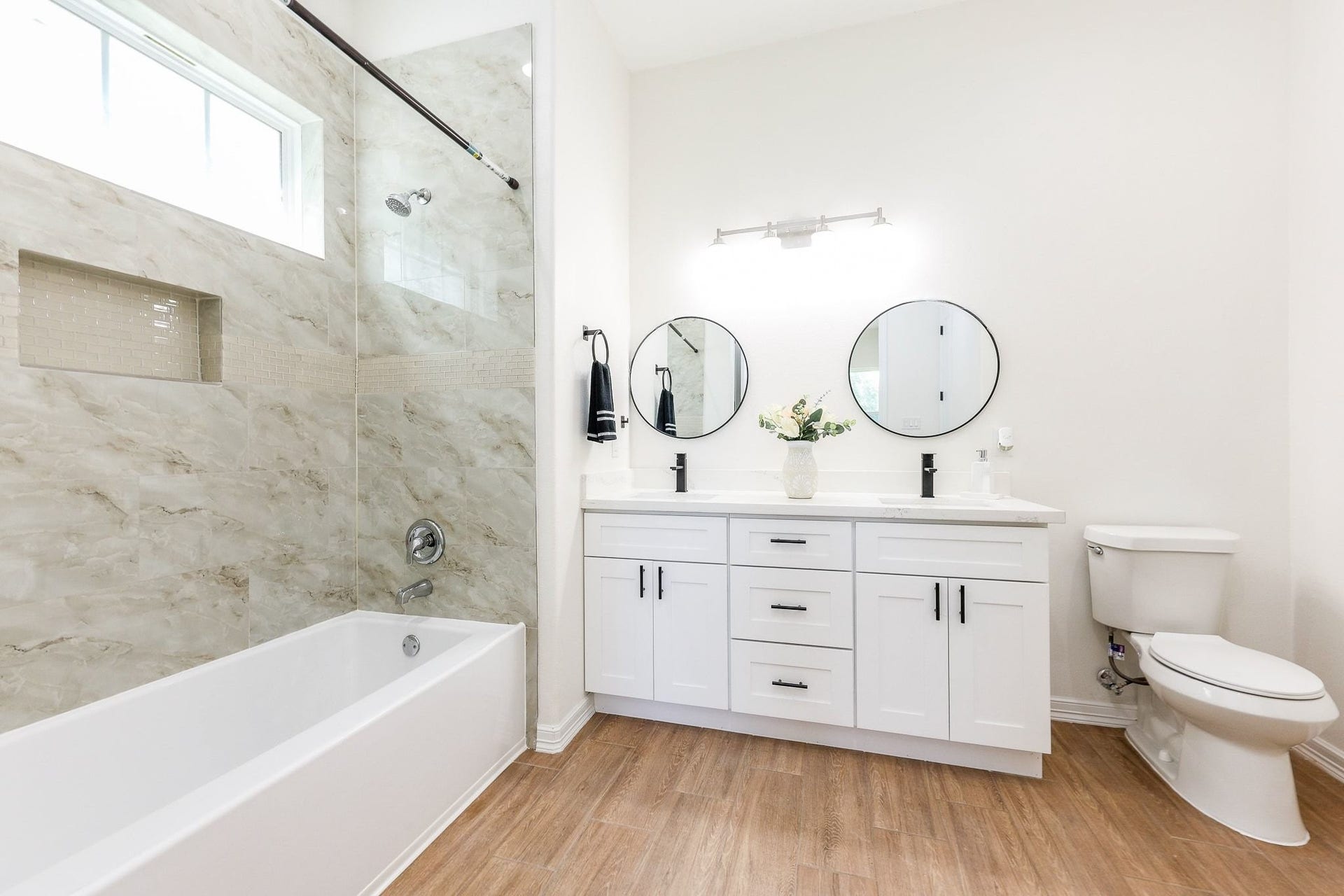 Bright and modern bathroom featuring a sleek shower, elegant double vanity with round mirrors, and warm wood-style flooring.