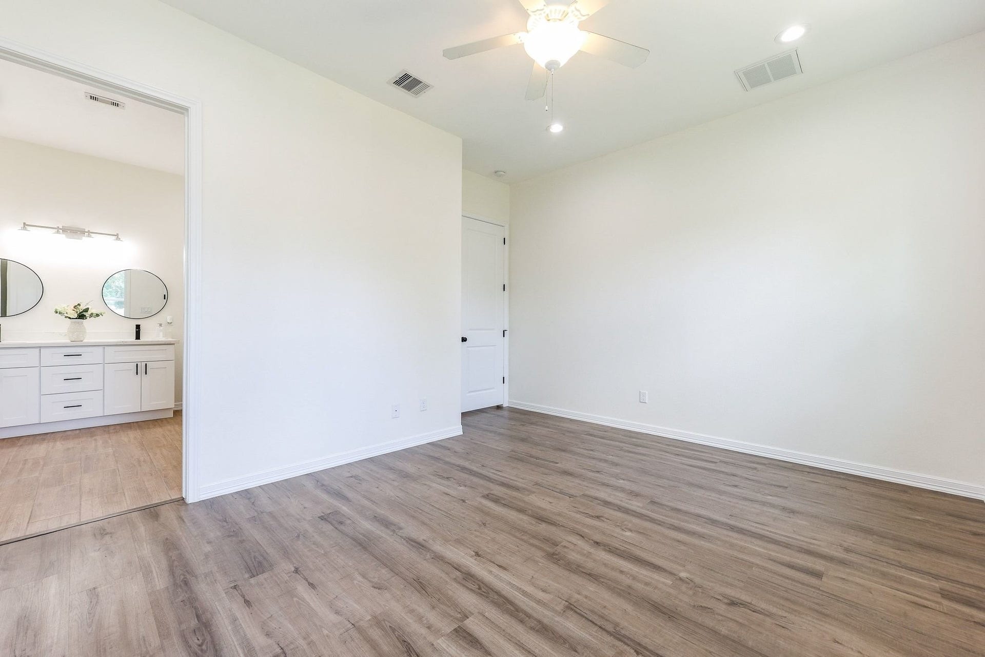 Spacious room with elegant laminate flooring and a ceiling fan, leading into a modern bathroom with dual sinks and stylish round mirrors.