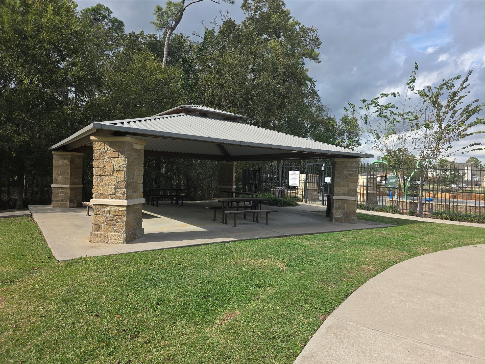 Charming outdoor pavilion with stone pillars and a metal roof, perfect for gatherings, surrounded by lush greenery and located near recreational facilities.