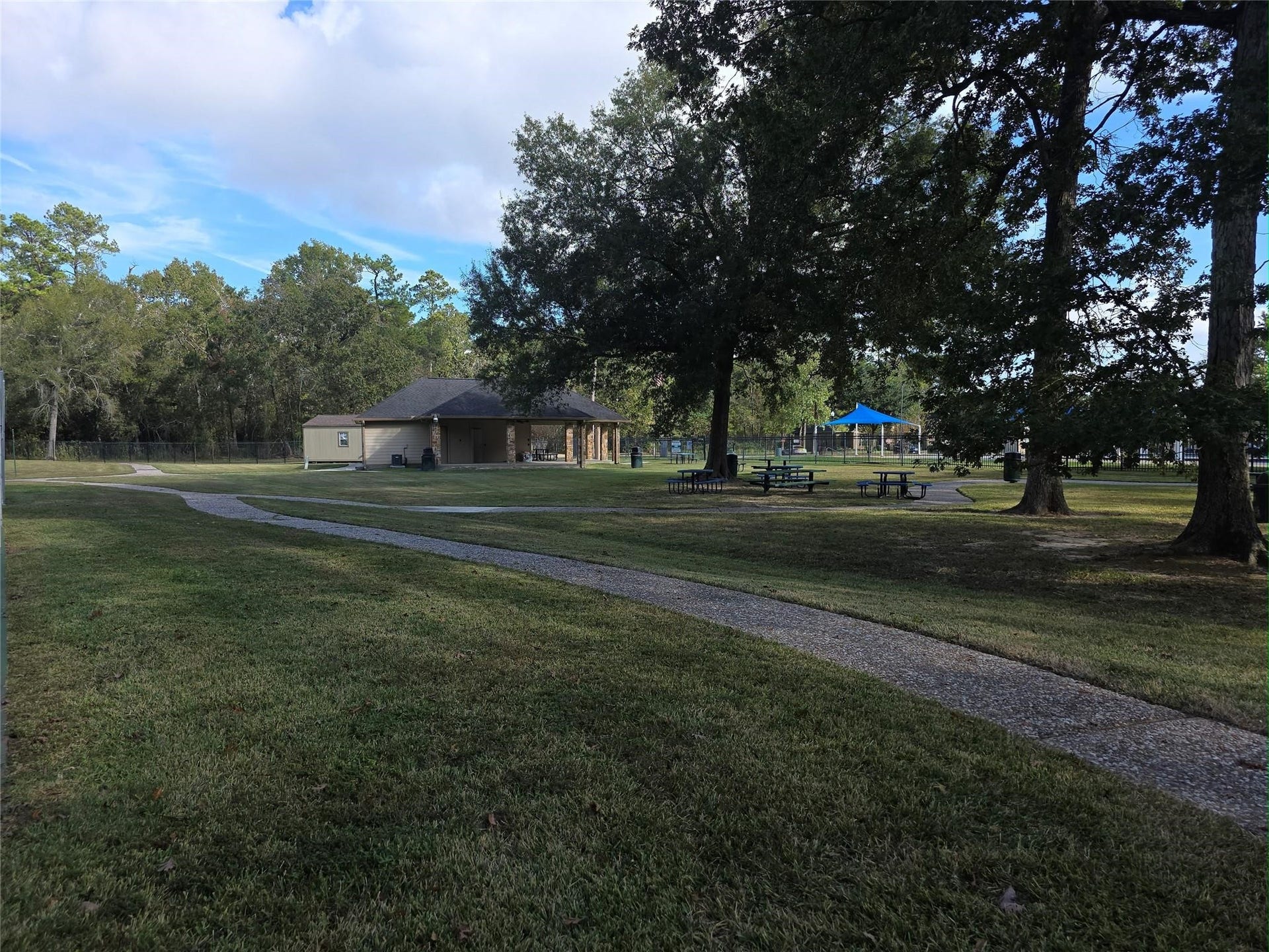 Expansive green space with a winding path leads to a cozy pavilion and picnic area, surrounded by mature trees and a peaceful natural setting.