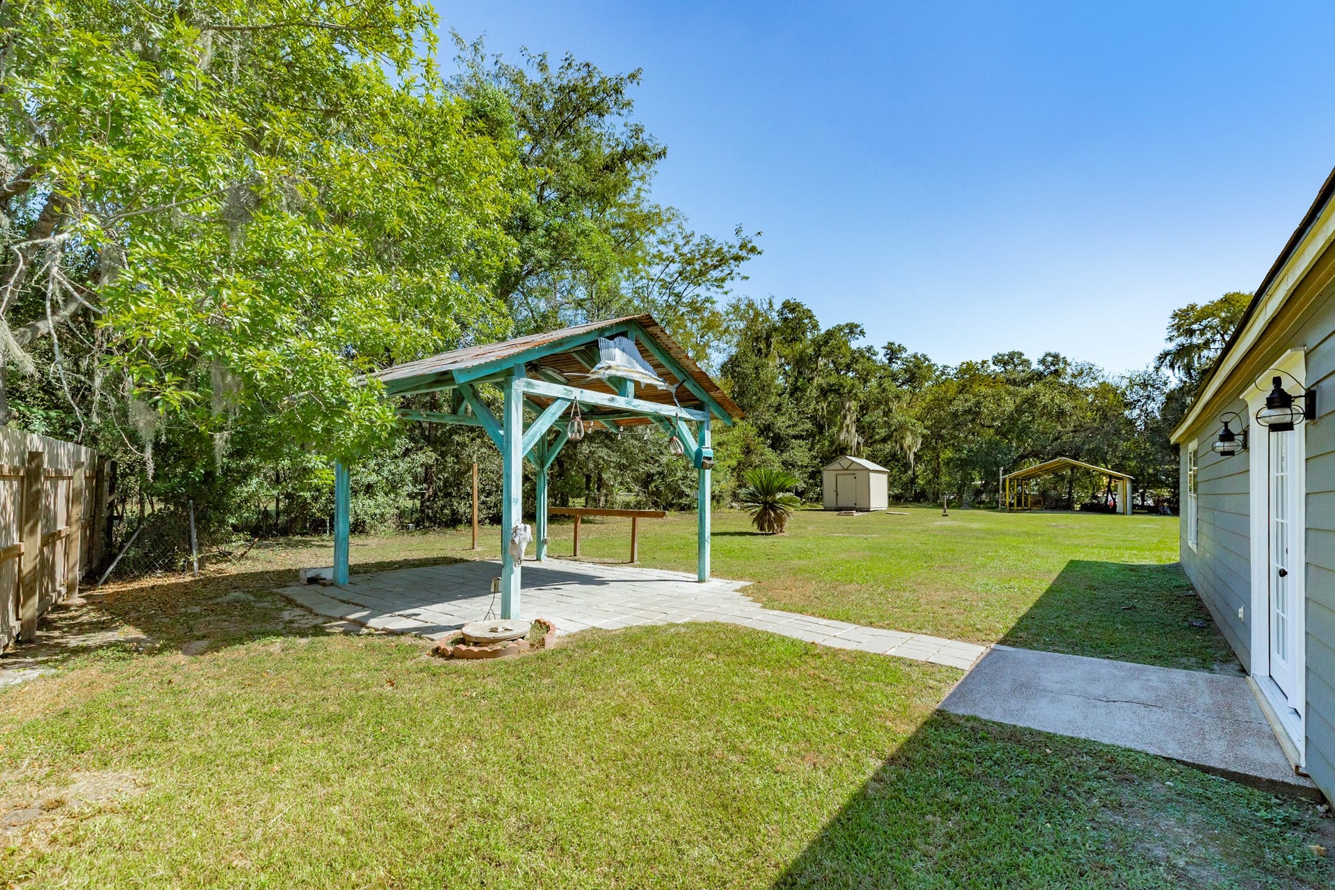 1602 County Road 939, Sweeny, TX 77480 Spacious backyard offers a covered pavilion connected by a paved walkway, surrounded by mature shade trees and open lawn. Additional outdoor structures provide convenient options for storage and covered use.