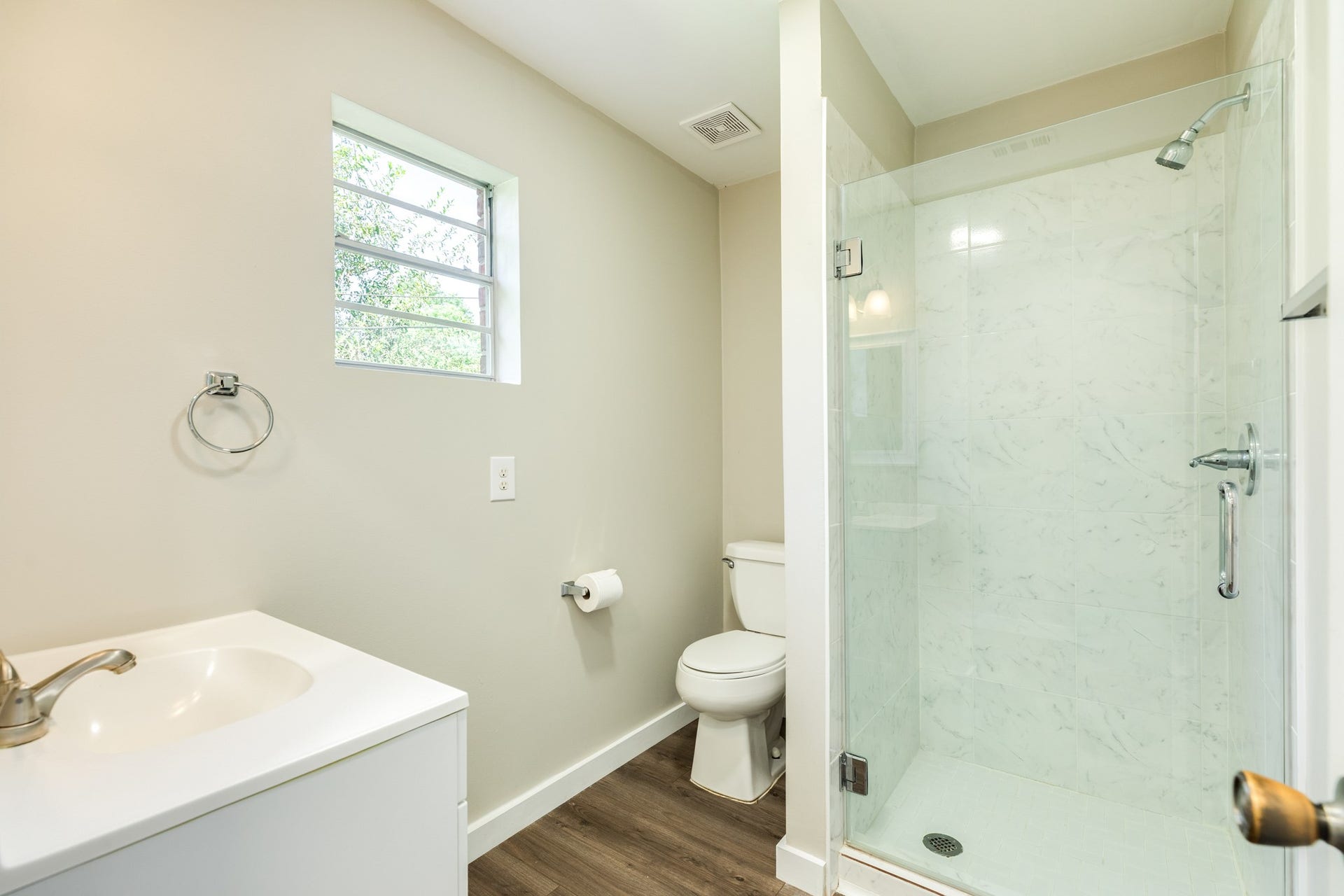 1602 County Road 939, Sweeny, TX 77480 Fresh, neutral bathroom features a glass-enclosed walk-in shower with marble-look wall tile, a white vanity, and wood-look flooring. A window provides natural light and ventilation.