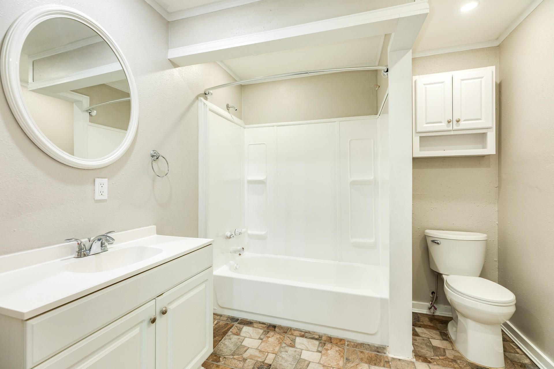 1602 County Road 939, Sweeny, TX 77480 Full bathroom features a white vanity, oval mirror, tub and shower combination with built-in shelving, and additional wall-mounted cabinetry. Decorative stone-look flooring adds warmth and character.