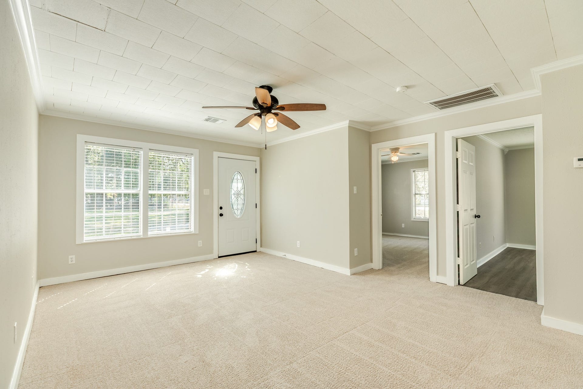1602 County Road 939, Sweeny, TX 77480 Bright and inviting living room features neutral carpeting, soft wall colors, decorative ceiling panels, and a ceiling fan with lighting. Large windows bring in natural light, while the decorative glass entry door adds character.