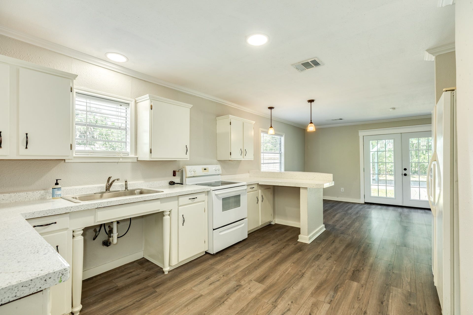 1602 County Road 939, Sweeny, TX 77480 Spacious kitchen features white cabinetry, speckled countertops, a double-basin sink, and a breakfast bar overlooking the dining area. Wood-look flooring, pendant lighting, and French doors create an inviting space for everyday living and entertaining.