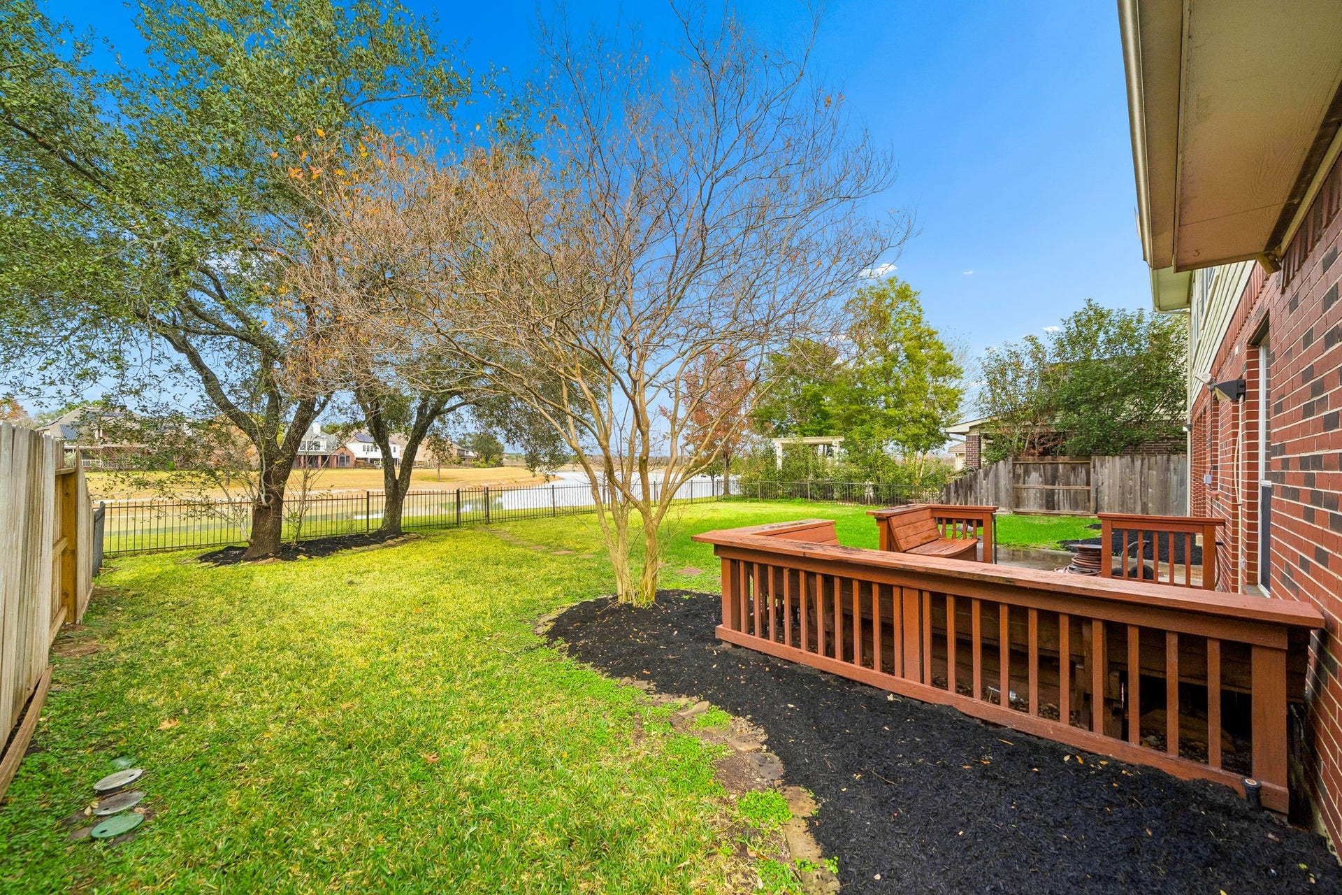 This photo showcases a backyard with a wooden deck attached to a brick house. The green lawn is bordered by trees and a fence, offering a serene view of a nearby pond or lake. Ideal for relaxation and outdoor activities.