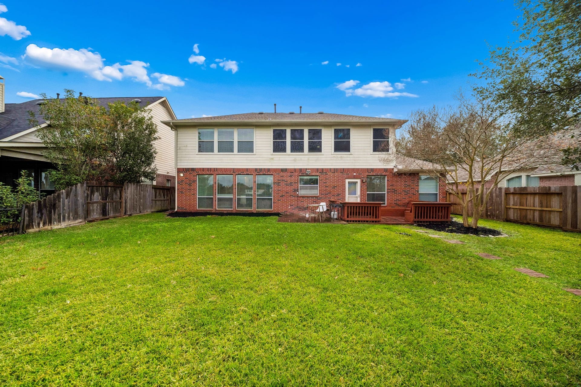 Spacious two-story brick and siding home with a large, well-maintained backyard. Features a small deck area, mature trees, and a wooden fence for privacy.