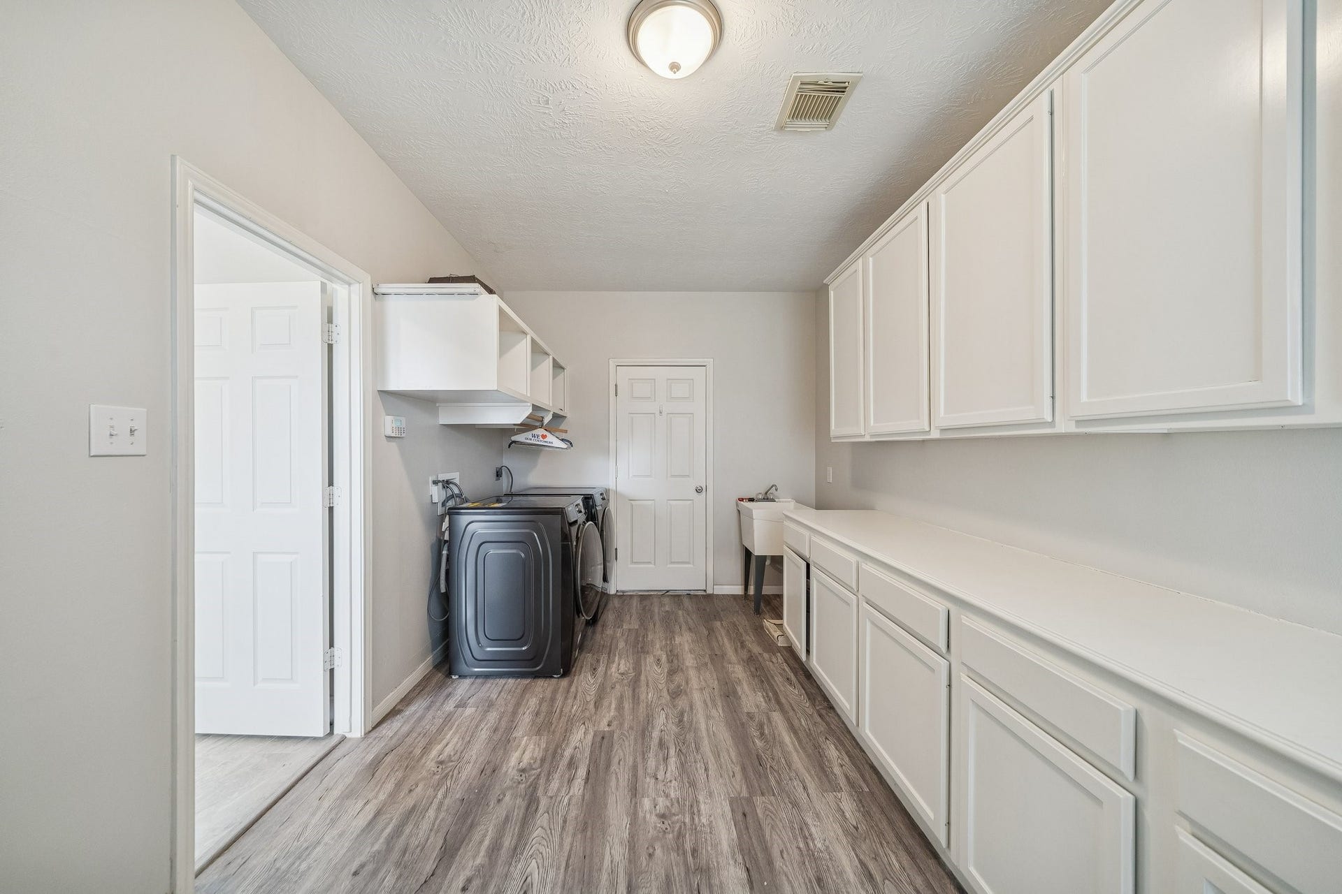 Bright and spacious laundry room featuring ample white cabinetry, modern washer and dryer, a utility sink, and sleek wood-style flooring.