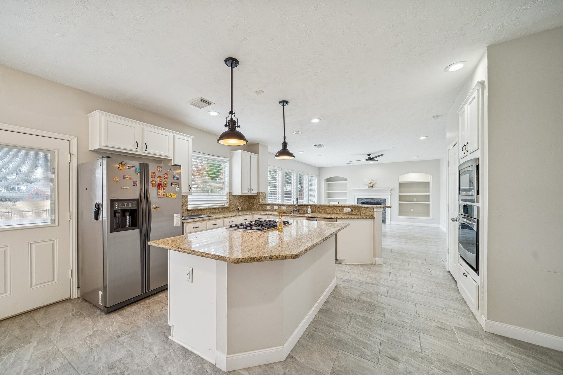 This spacious kitchen features modern appliances, a large granite island with a cooktop, and ample natural light. It opens to a bright living area with built-in shelving and a cozy fireplace, offering a welcoming and functional space for family gatherings.