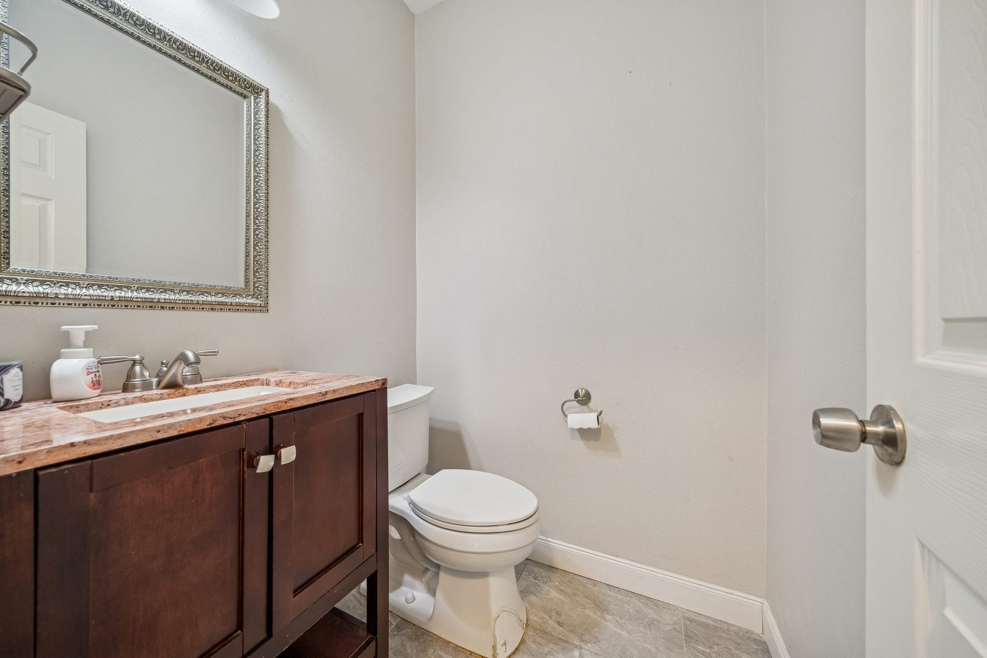 This is a modern half-bathroom featuring a sleek vanity with a marble countertop, a large decorative mirror, and a simple toilet, all set against neutral walls and tile flooring.