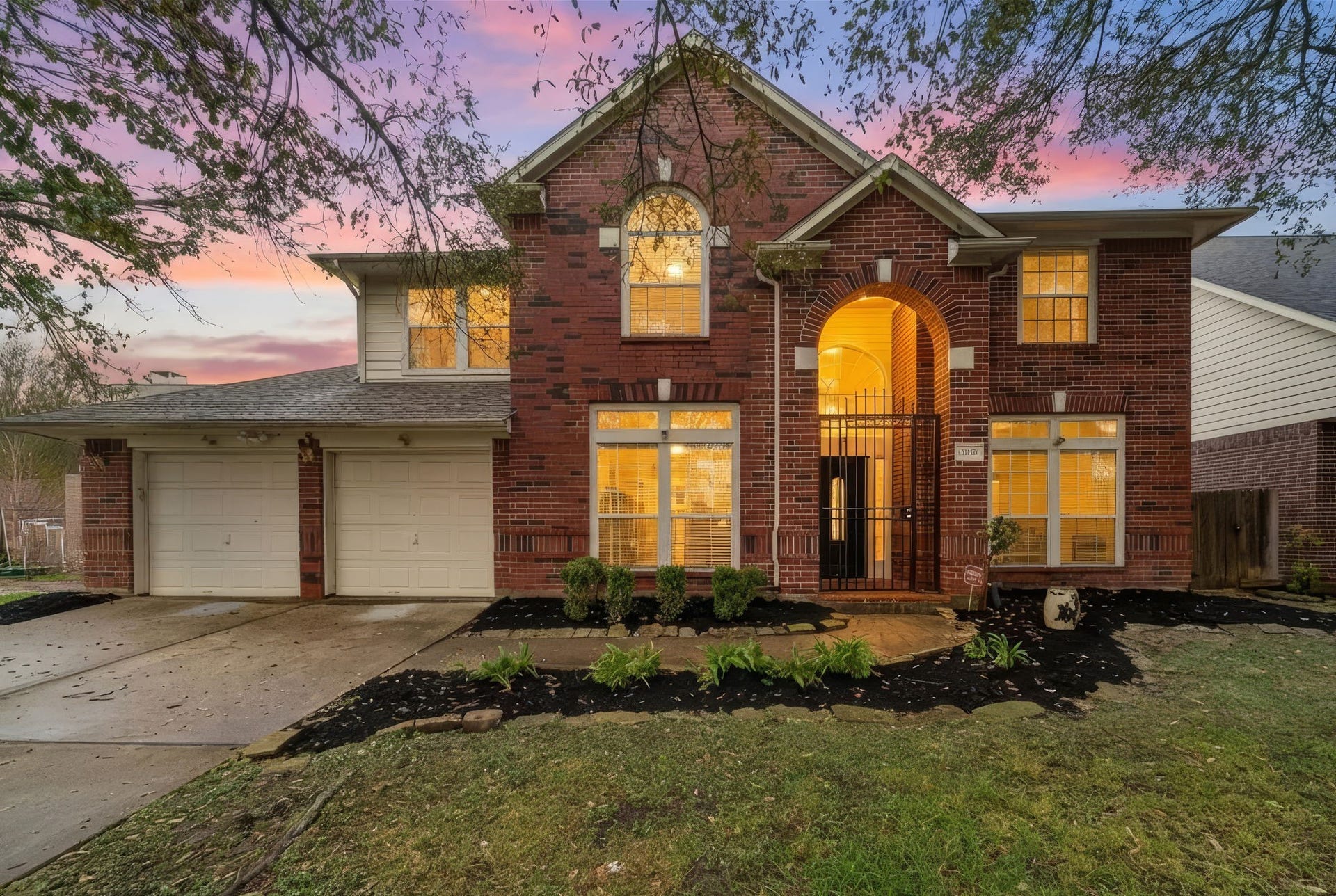 This stunning two-story brick home features a welcoming arched entryway, large windows, and a two-car garage. The beautifully landscaped front yard complements the warm, inviting interior lighting, all set against a picturesque sunset backdrop.