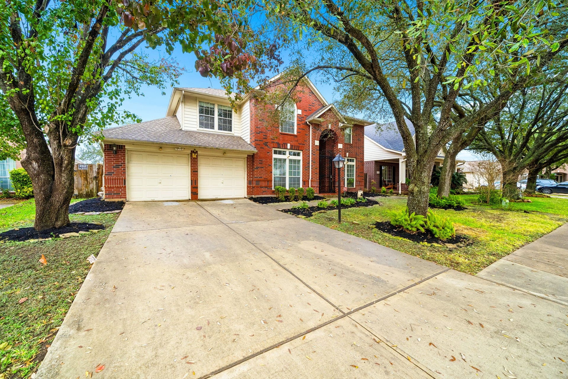 This charming home features a two-car garage and a brick exterior, nestled among mature trees. The front yard is well-maintained, with a spacious driveway and inviting entrance.