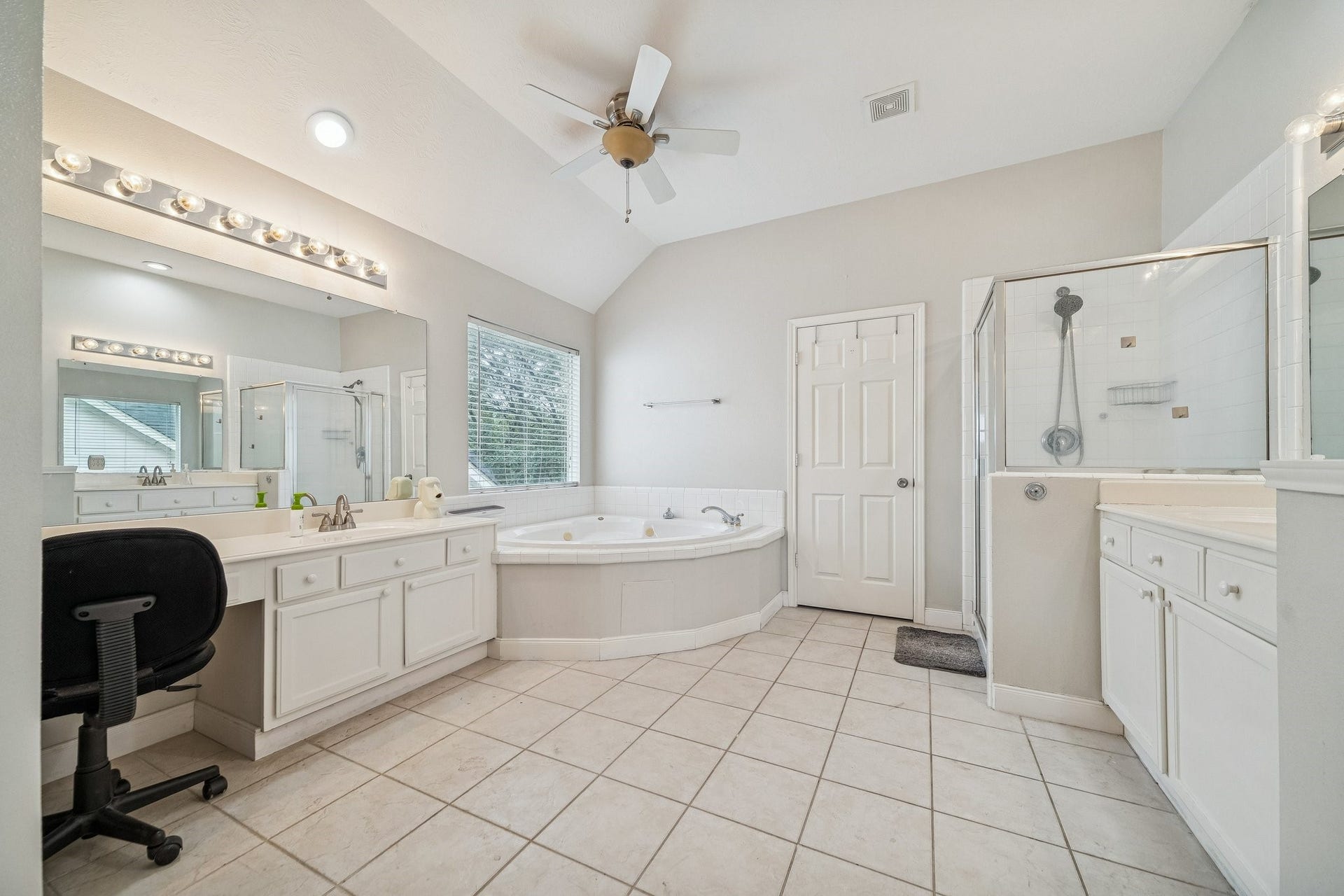 This spacious bathroom features a large corner bathtub, a separate glass-enclosed shower, dual sinks with ample counter space, and a ceiling fan. The neutral tile and ample natural light create a bright, inviting atmosphere.
