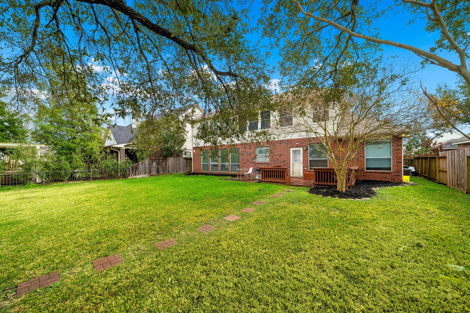 This photo showcases a spacious backyard with a well-maintained lawn and a brick pathway leading to a two-story brick house. The area is lined with mature trees, providing shade and privacy, ideal for outdoor activities and relaxation.
