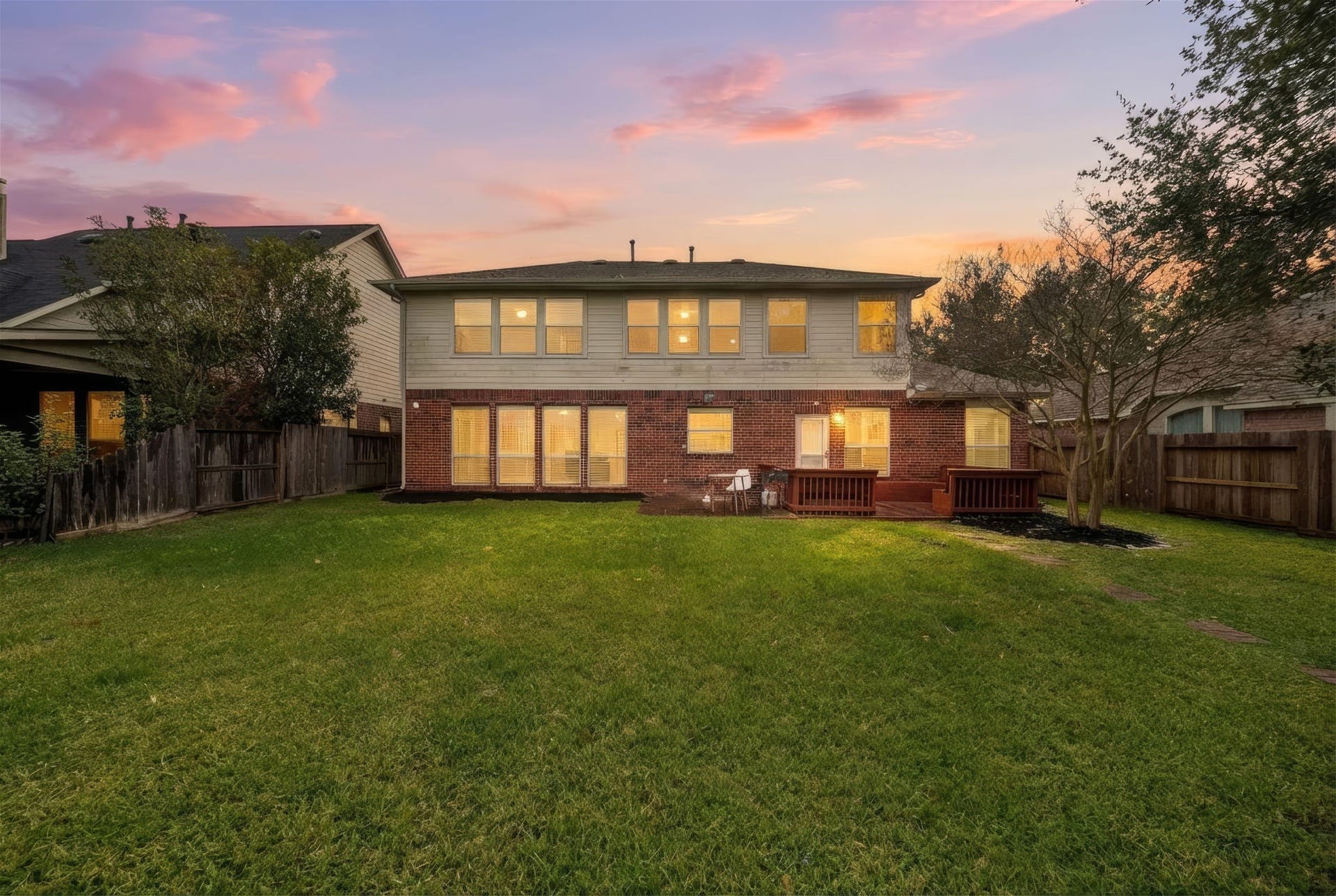This photo showcases the backyard of a two-story home with a spacious lawn, a wooden deck, and a cozy fence. The exterior features a combination of brick and siding, and the warm glow from the windows suggests a welcoming interior. The sky is beautifully lit at sunset, adding to the property's charm.