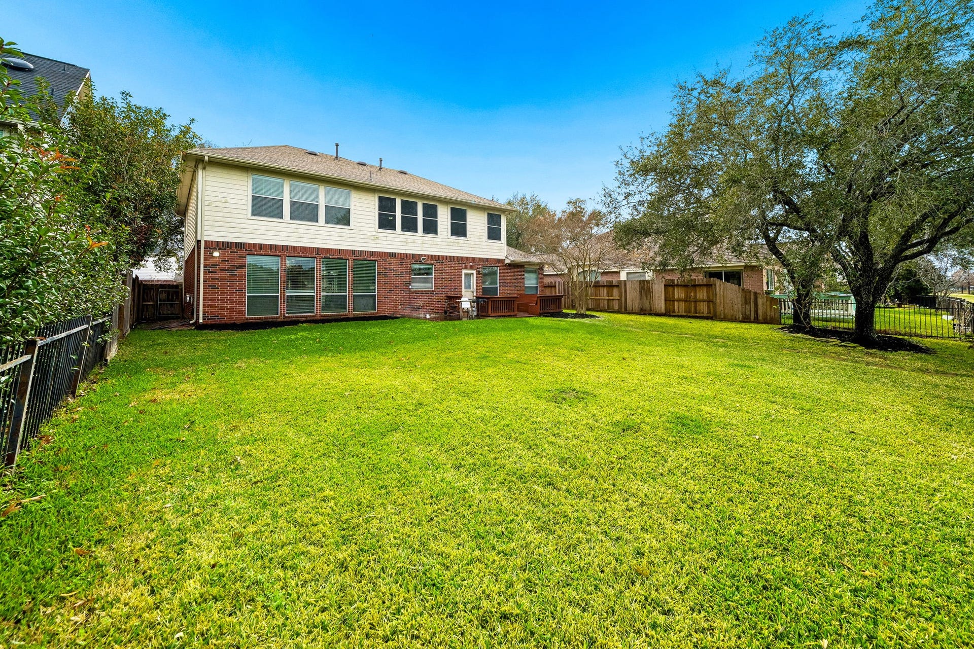 Spacious backyard with lush green lawn and mature trees, perfect for outdoor activities. The two-story brick and siding house features large windows and a small deck area, providing a great space for relaxation and entertainment.