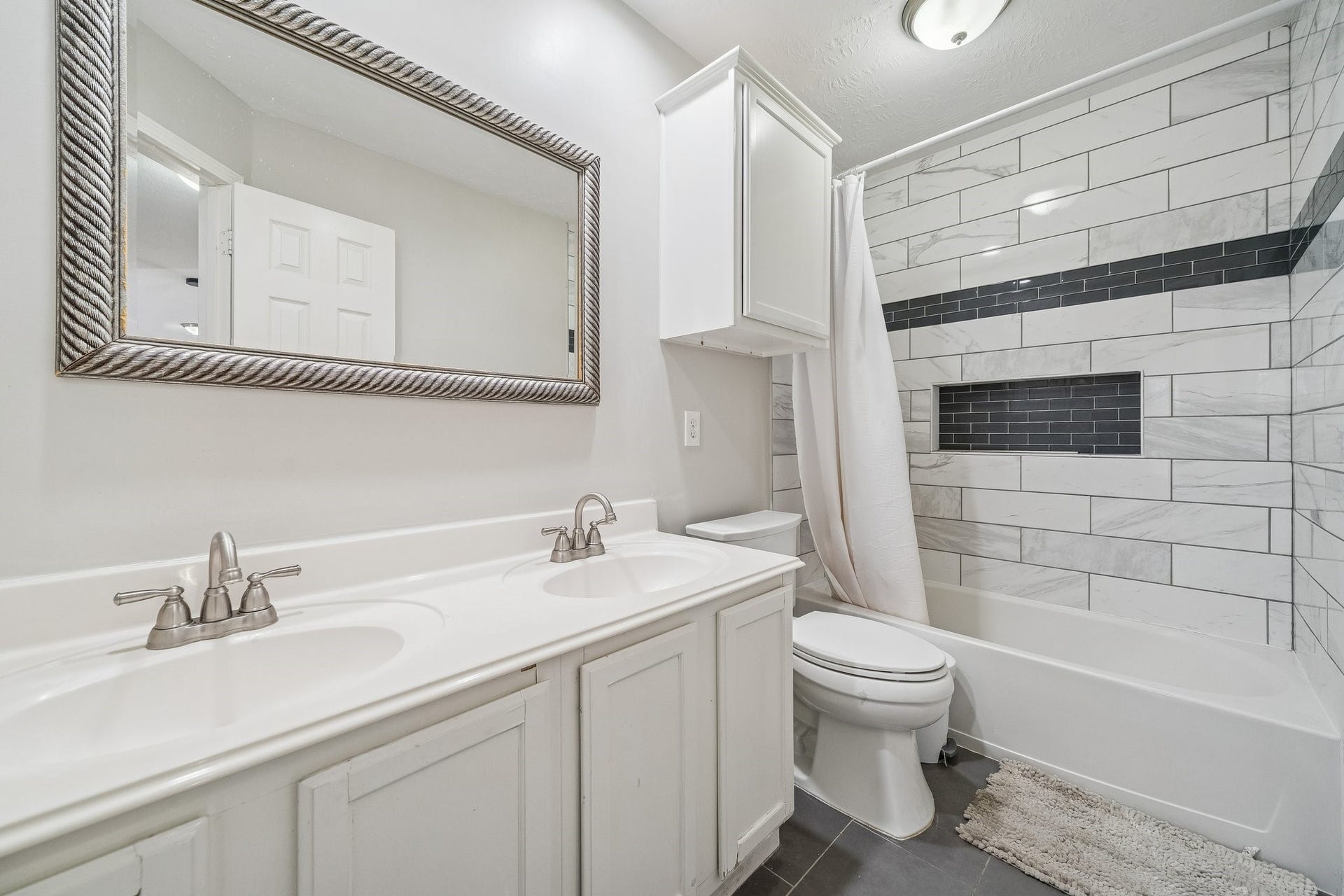 This bathroom features a modern design with a double sink vanity, large framed mirror, and a shower-tub combo with stylish tile work. Ample lighting and neutral tones create a bright, inviting space.