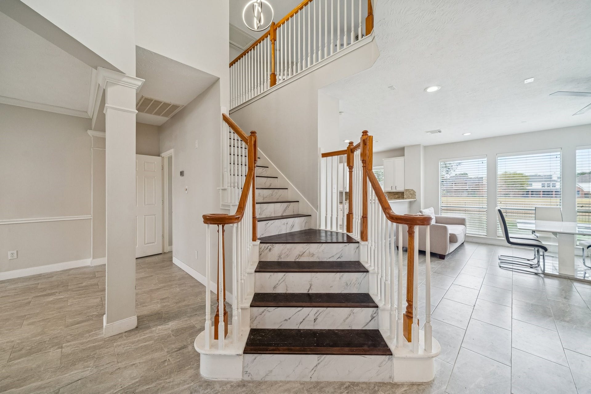 This photo showcases a bright, modern entryway with a stylish staircase featuring wooden handrails and marble steps. The open floor plan includes a living area with large windows, allowing plenty of natural light. The space is airy and inviting, perfect for family gatherings.