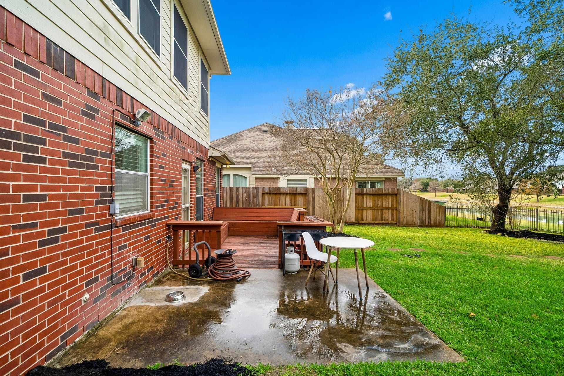 This photo showcases a spacious backyard with a brick exterior, a small patio area with seating, and a wooden deck. The yard is fenced, has well-maintained grass, and is complemented by mature trees, offering a cozy outdoor space for relaxation and entertaining.