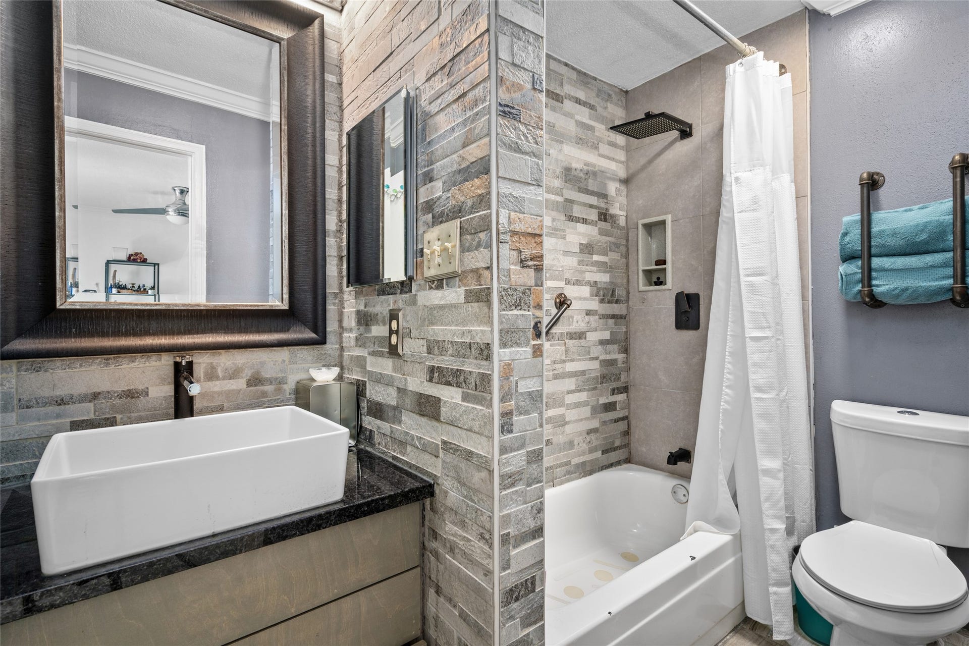 9850 Pagewood Lane, #1302, Houston, TX 77042 This beautifully updated primary bathroom features a modern, spa-like aesthetic with a striking stone accent wall and a sleek black granite vanity. The vessel sink and framed mirror add a touch of sophistication, while the walk-in shower boasts a rainfall showerhead for a luxurious bathing experience. A deep soaking tub provides an additional option for relaxation. Thoughtful storage solutions, including built-in shelving and industrial-style towel racks, enhance both style and functionality.