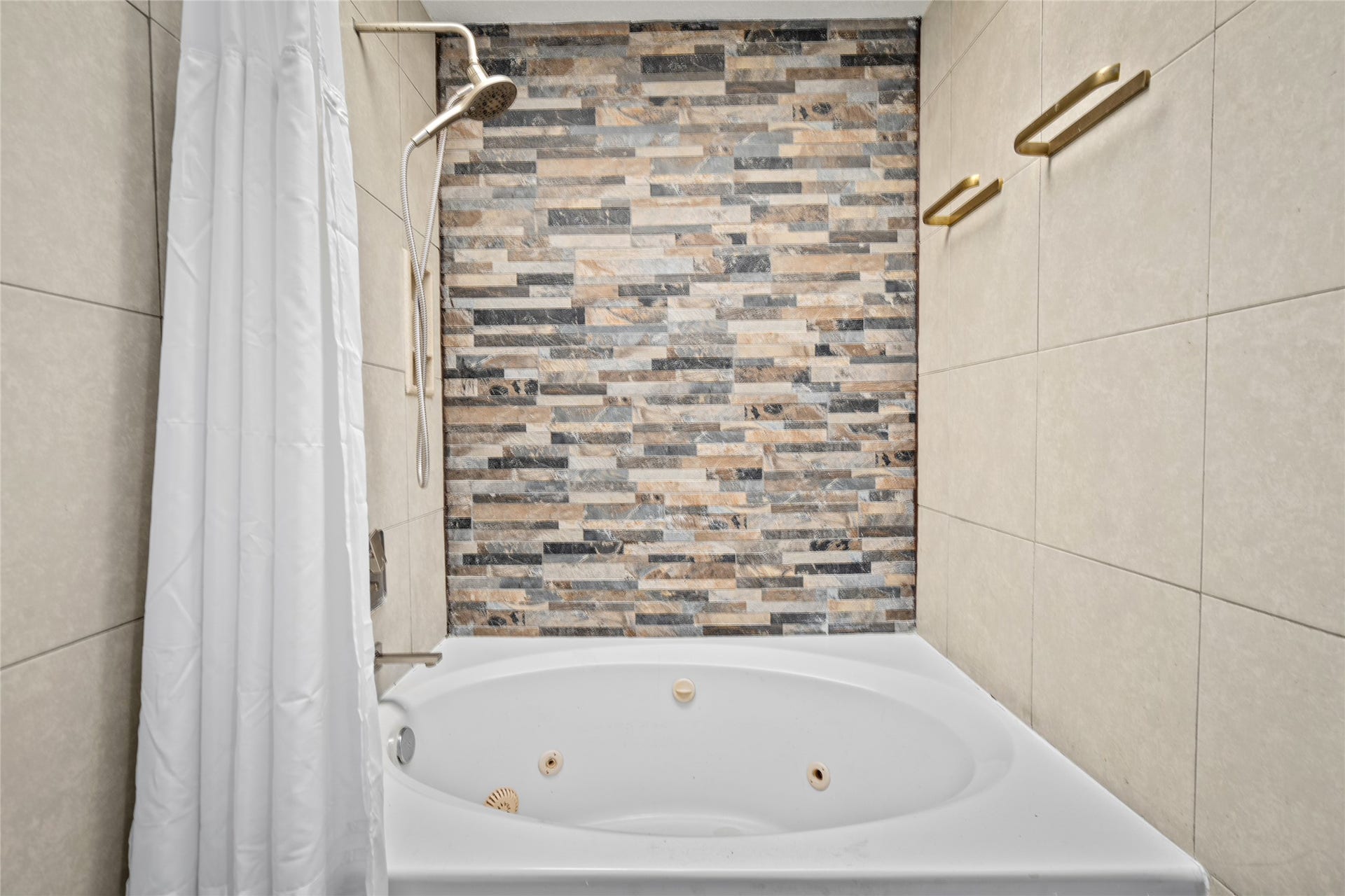 9850 Pagewood Lane, #1302, Houston, TX 77042 Unwind in the luxurious primary bath featuring a relaxing jetted tub, perfect for soaking after a long day. The stunning accent wall with modern stacked stone tile adds a touch of elegance, while the handheld showerhead provides both convenience and style. Sleek, neutral-toned tiles and gold-tone towel racks complete this spa-like retreat.