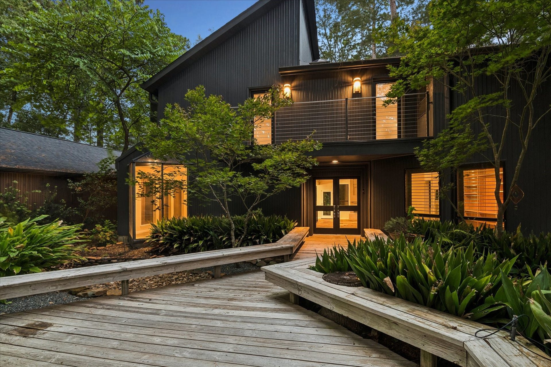 23414 Holly Hollow Street, Tomball, TX 77377 A striking blue-hour arrival: the home's dark vertical siding and soaring gable glow warmly from within, framed by mature trees and a Japanese maple. A wraparound wood boardwalk winds through lush landscaping to the double-door entry, while the cable-railed upper balcony hints at the architectural drama inside.
