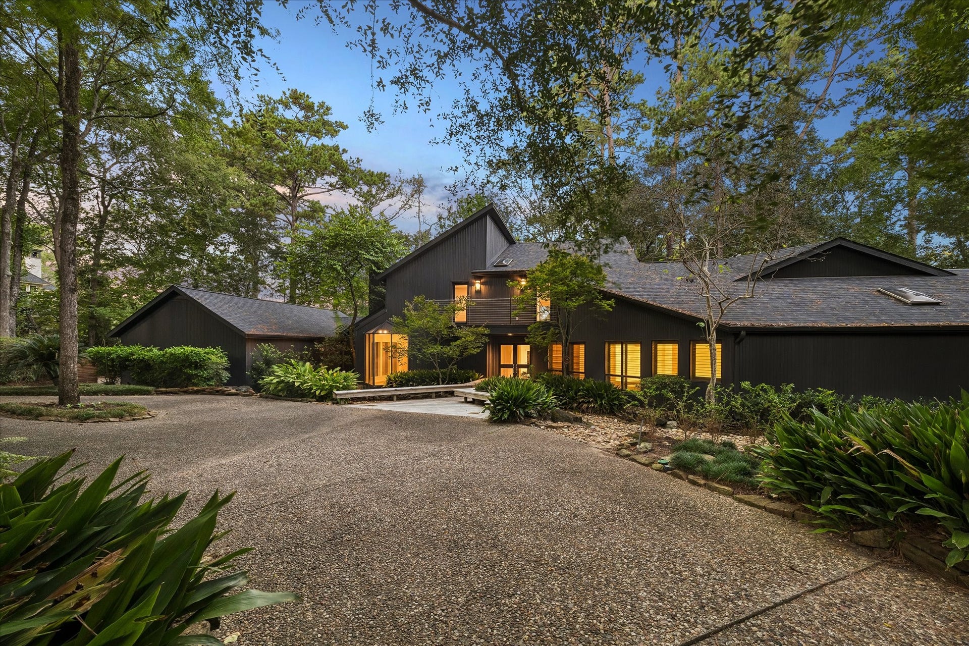 23414 Holly Hollow Street, Tomball, TX 77377 A sweeping gravel drive curves through towering pines and lush understory plantings toward this striking modern home. Multiple gable rooflines and board-formed siding create bold architectural silhouettes against the evening sky, while a wooden boardwalk guides guests to the illuminated entrance total privacy just steps from the front door.