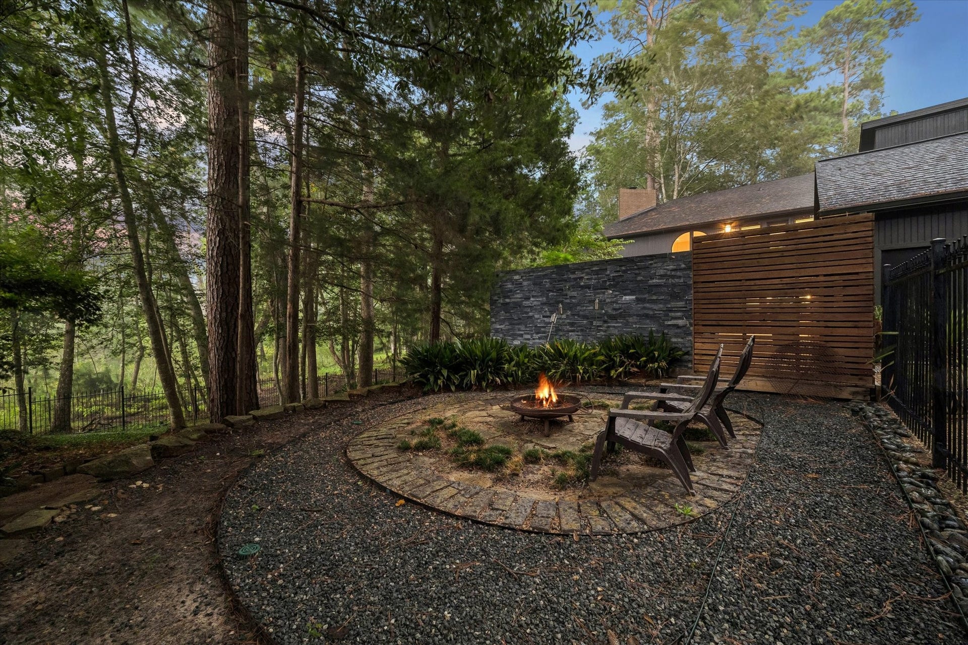 23414 Holly Hollow Street, Tomball, TX 77377 Tucked into the wooded backyard, a circular stone fire pit patio offers an intimate gathering spot surrounded by native plantings and a dramatic slate stone wall. Adirondack chairs invite quiet evenings by the fire, with a sleek wood-slat privacy screen adding modern texture against the natural setting.