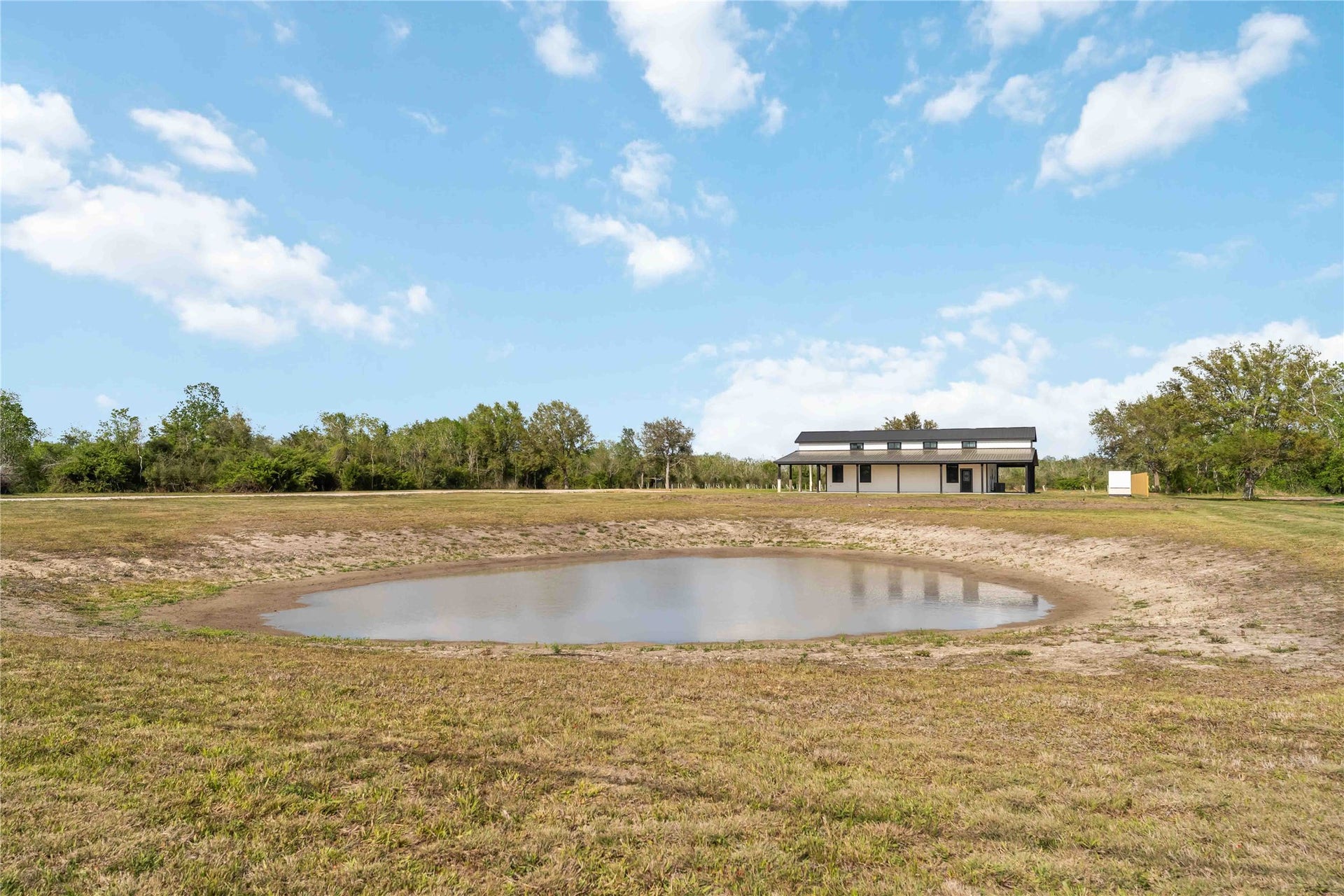 4864 County Road 182 Off, Alvin, TX 77511 A modern, single-story home is situated on a spacious plot with a small pond in the foreground. The surrounding area is open and grassy, bordered by trees under a clear blue sky.