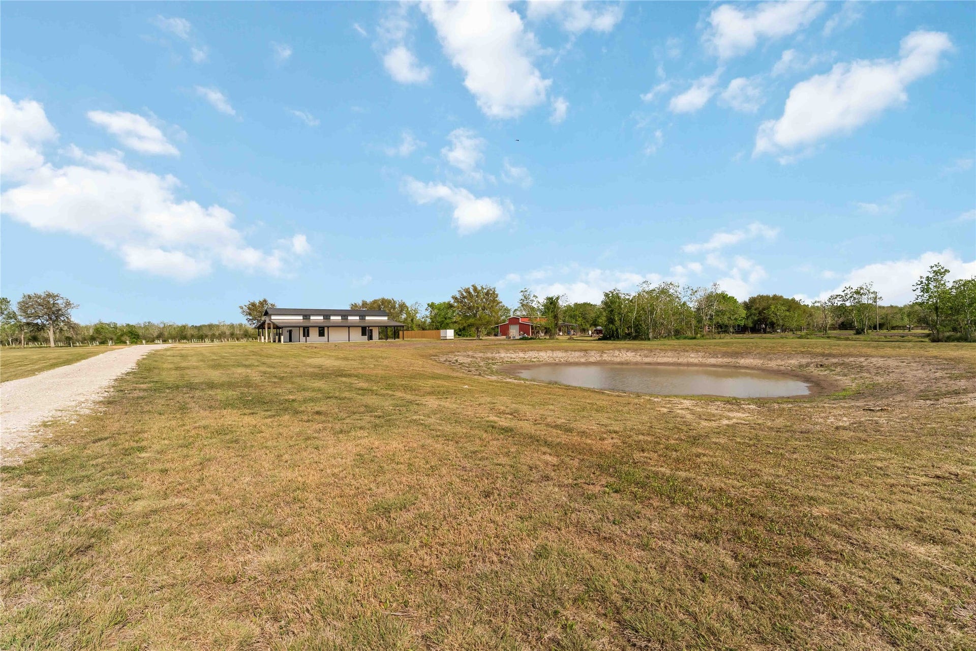 4864 County Road 182 Off, Alvin, TX 77511 Expansive property with a modern house, a small pond, and a long gravel driveway, surrounded by open land and trees, offering a serene rural setting.