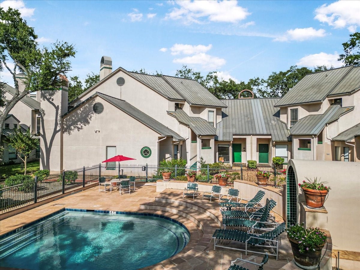 4144 Greystone Way, #101, Sugar Land, TX 77479 This property features a charming exterior with a metal roof and a shared courtyard. It includes a fenced-in pool area with lounge chairs and a patio table, complemented by lush greenery and potted plants, perfect for relaxation and entertaining.