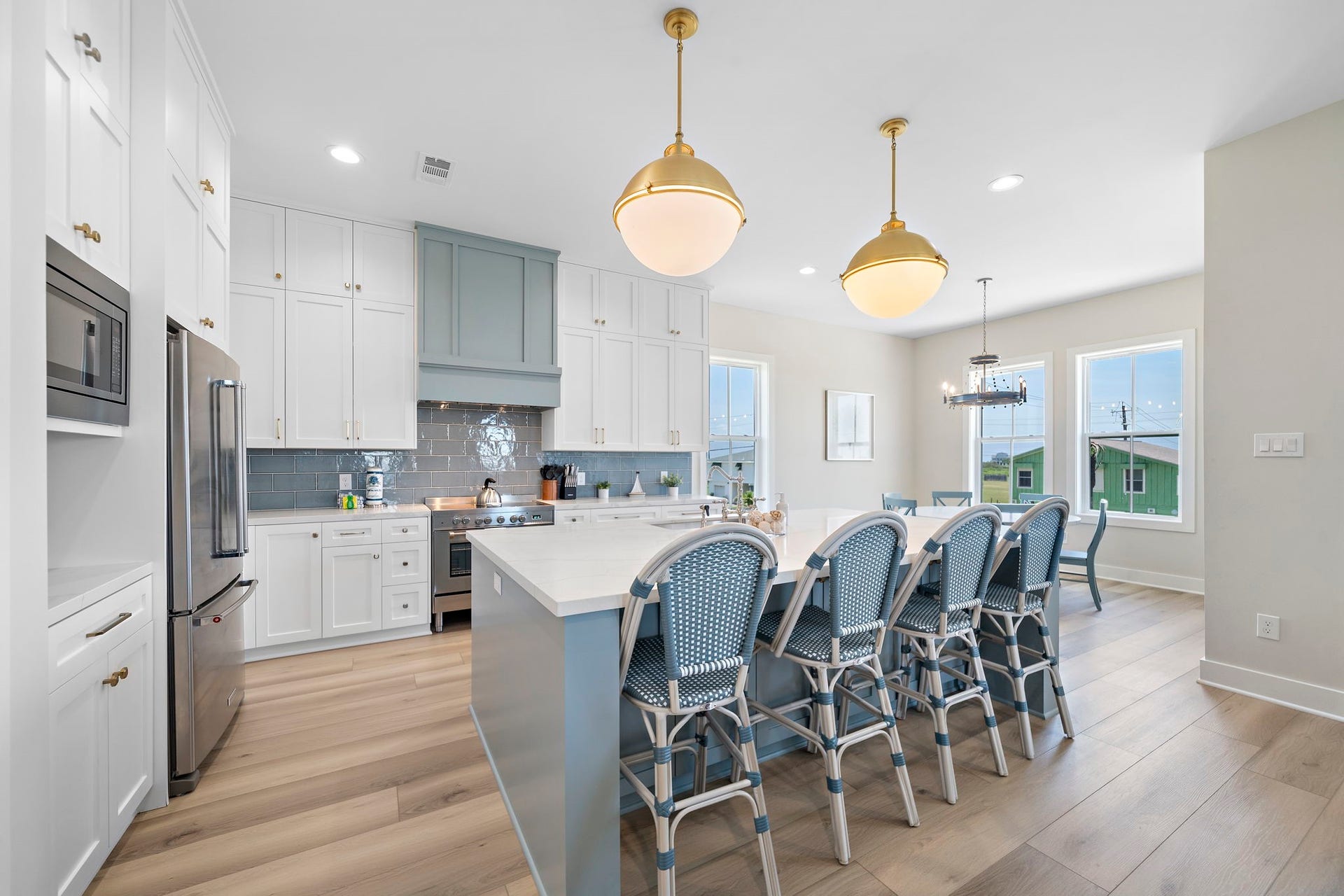 Bright and spacious kitchen featuring a large island with seating for six, elegant pendant lighting, and modern stainless steel appliances, complemented by crisp white cabinetry and a stylish blue tile backsplash.